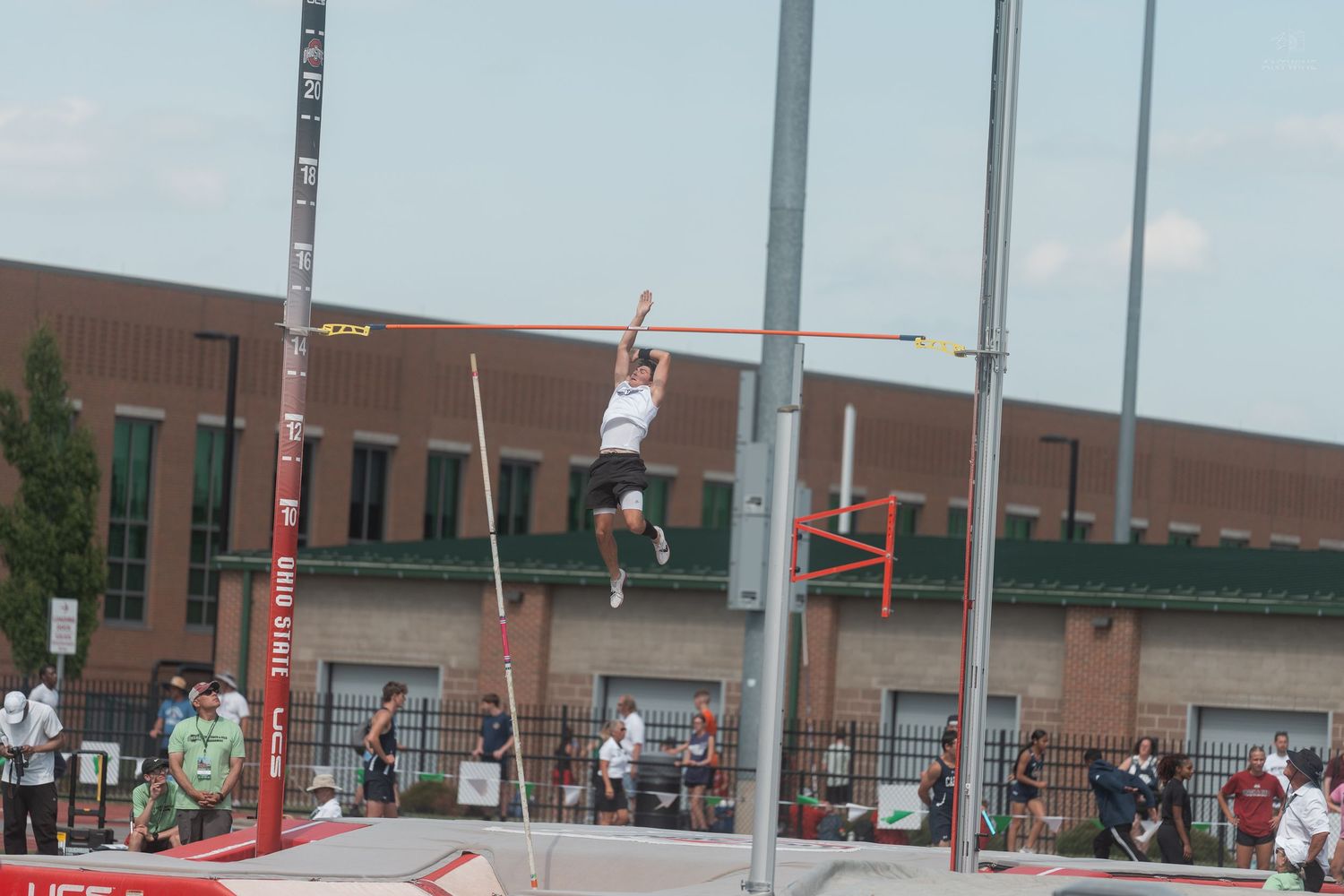 Young athlete ascends during a pole vault attempt at an outdoor track and field competition on a sunny day.