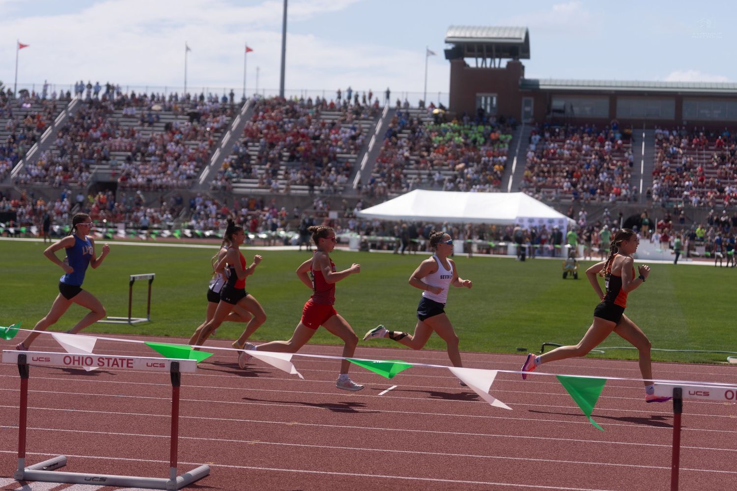 Female athletes competing in hurdles race at Ohio State track meet with packed stadium crowd.