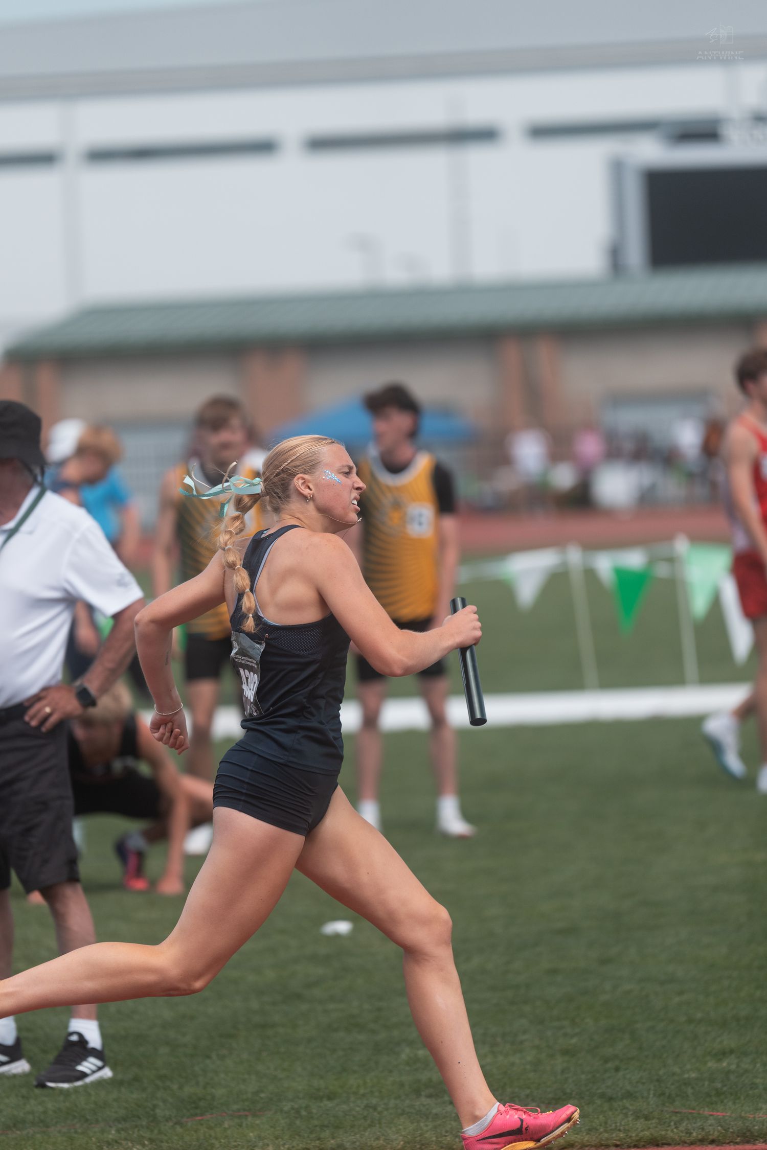 Female track athlete in black uniform runs relay leg holding baton at outdoor track meet.