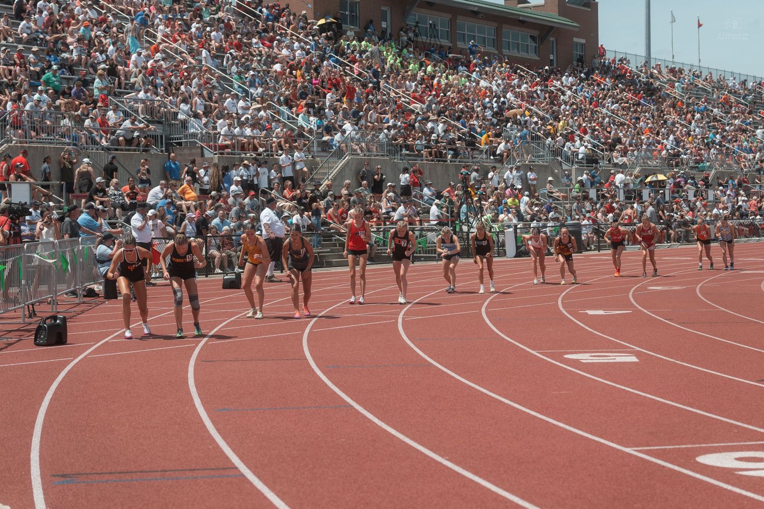 Female athletes run along the curved track during a high school track and field competition before a packed crowd.