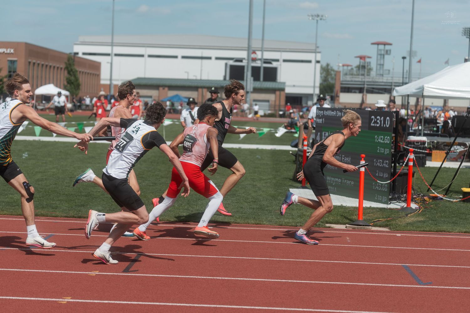High school track athletes compete in a relay race, passing batons on a red track at a sunny meet.