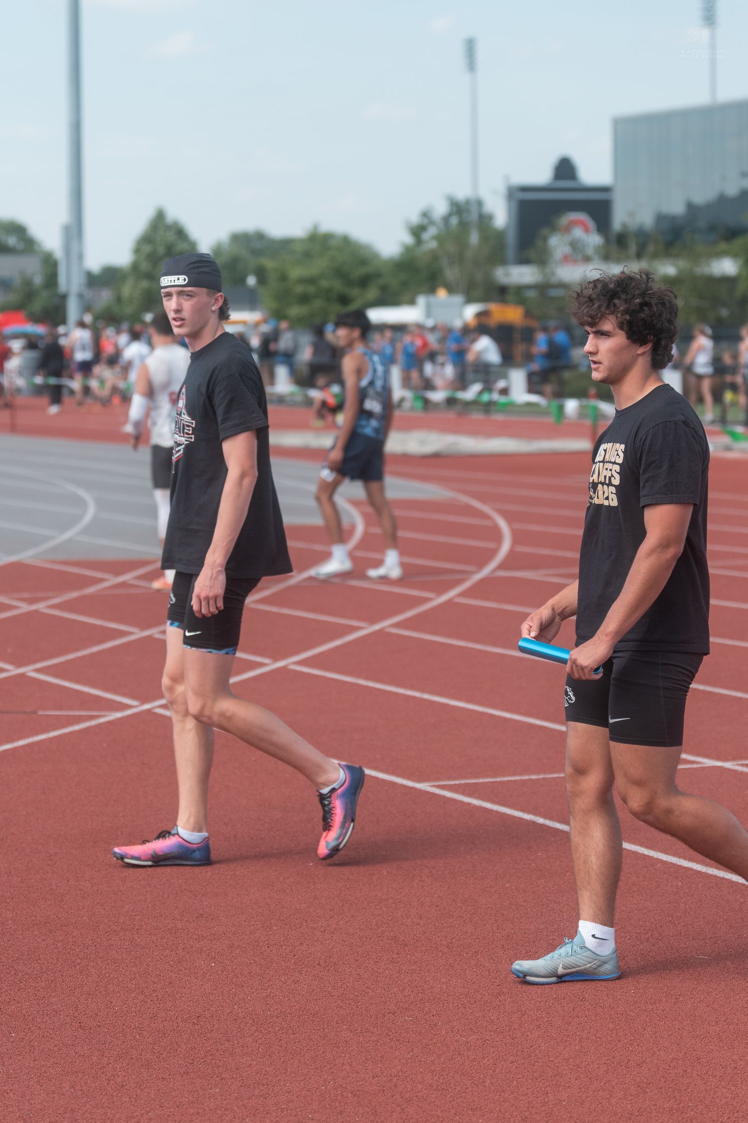 Two male track athletes in black uniforms walk on a red track during a busy outdoor relay competition.