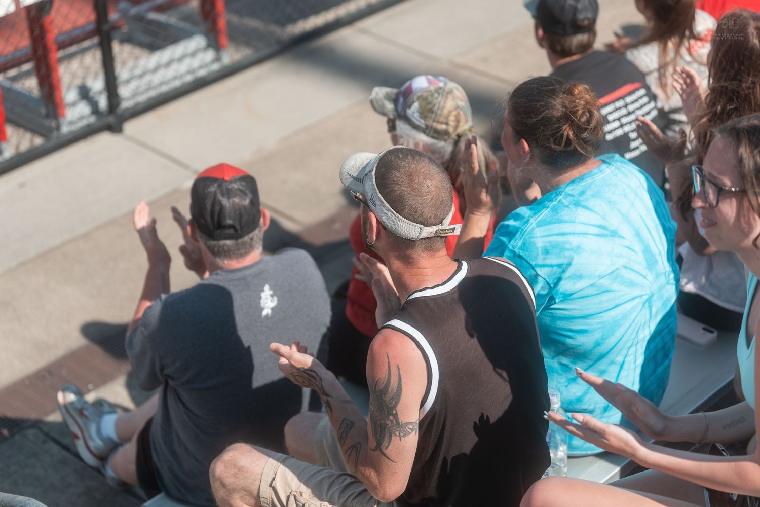 A group of spectators sit closely together watching an outdoor track and field competition on a sunny day.
