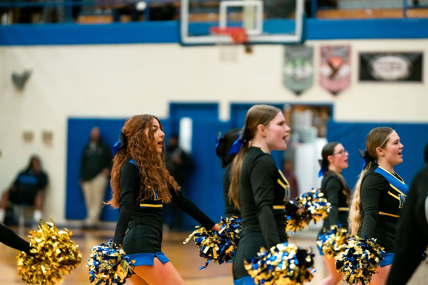 High school cheerleaders with gold and black pom-poms perform a routine on a gymnasium floor.