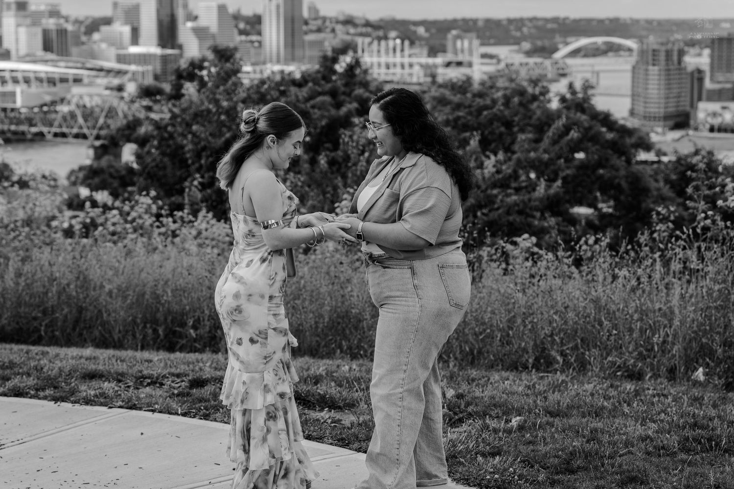 Two women holding hands sharing a romantic moment outdoors with city skyline backdrop in black and white.