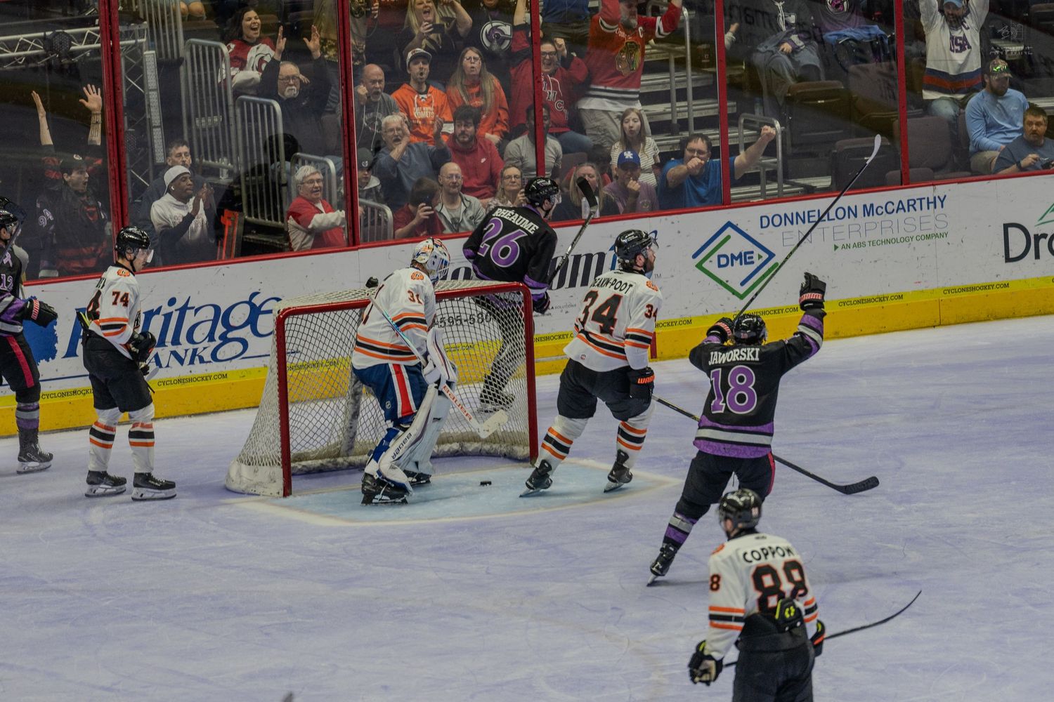 Hockey players celebrate a goal near the net during a competitive indoor ice hockey game with fans watching.
