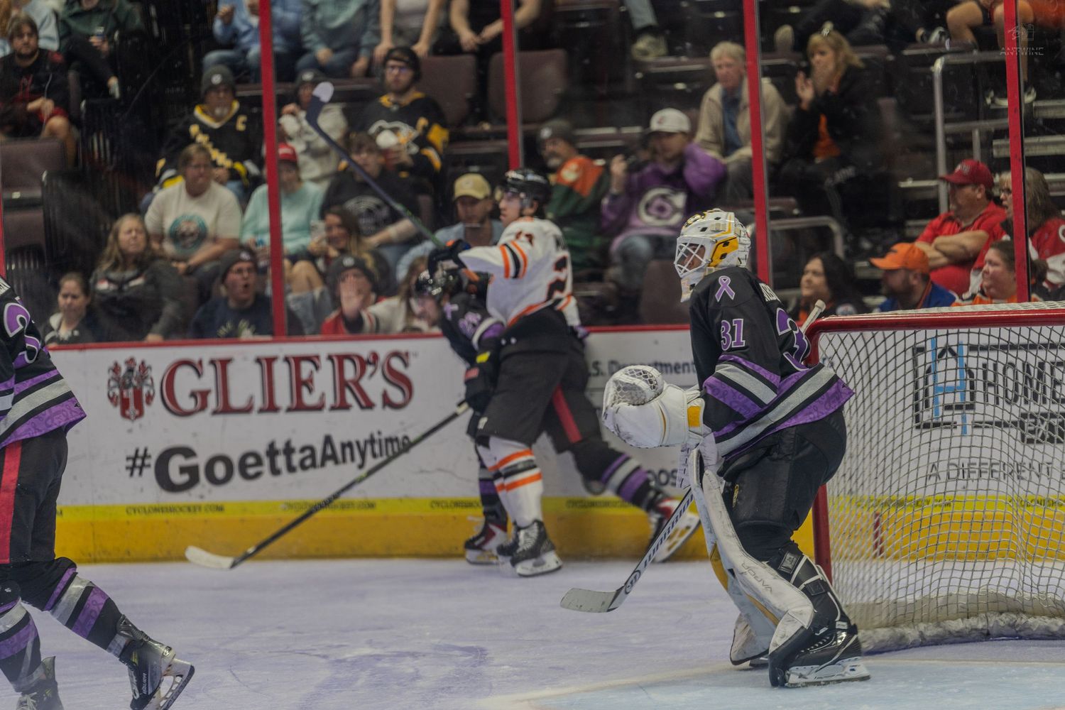 Orange-uniformed hockey player drives toward goal as purple-jersied goalie prepares to defend during ice hockey match.