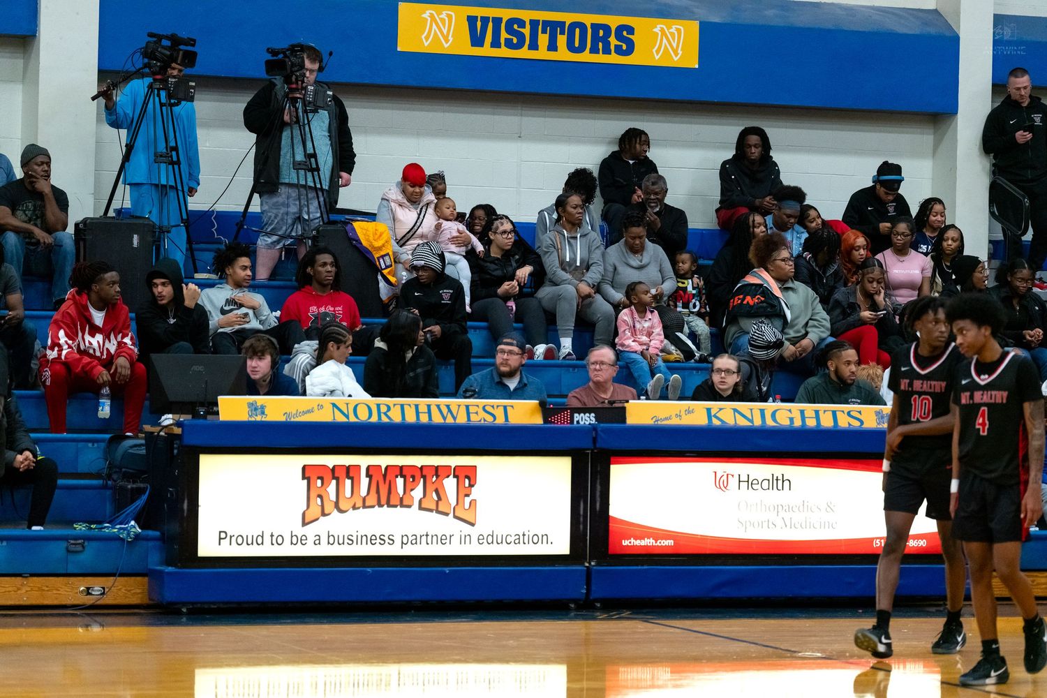 Crowded high school basketball game at Northwest Knights gymnasium with spectators and a TV cameraman in the stands.