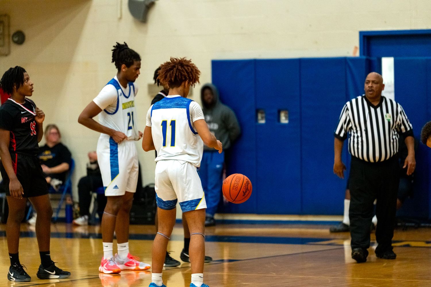 Two teammates in blue uniforms prepare for play as a referee approaches during a basketball game.