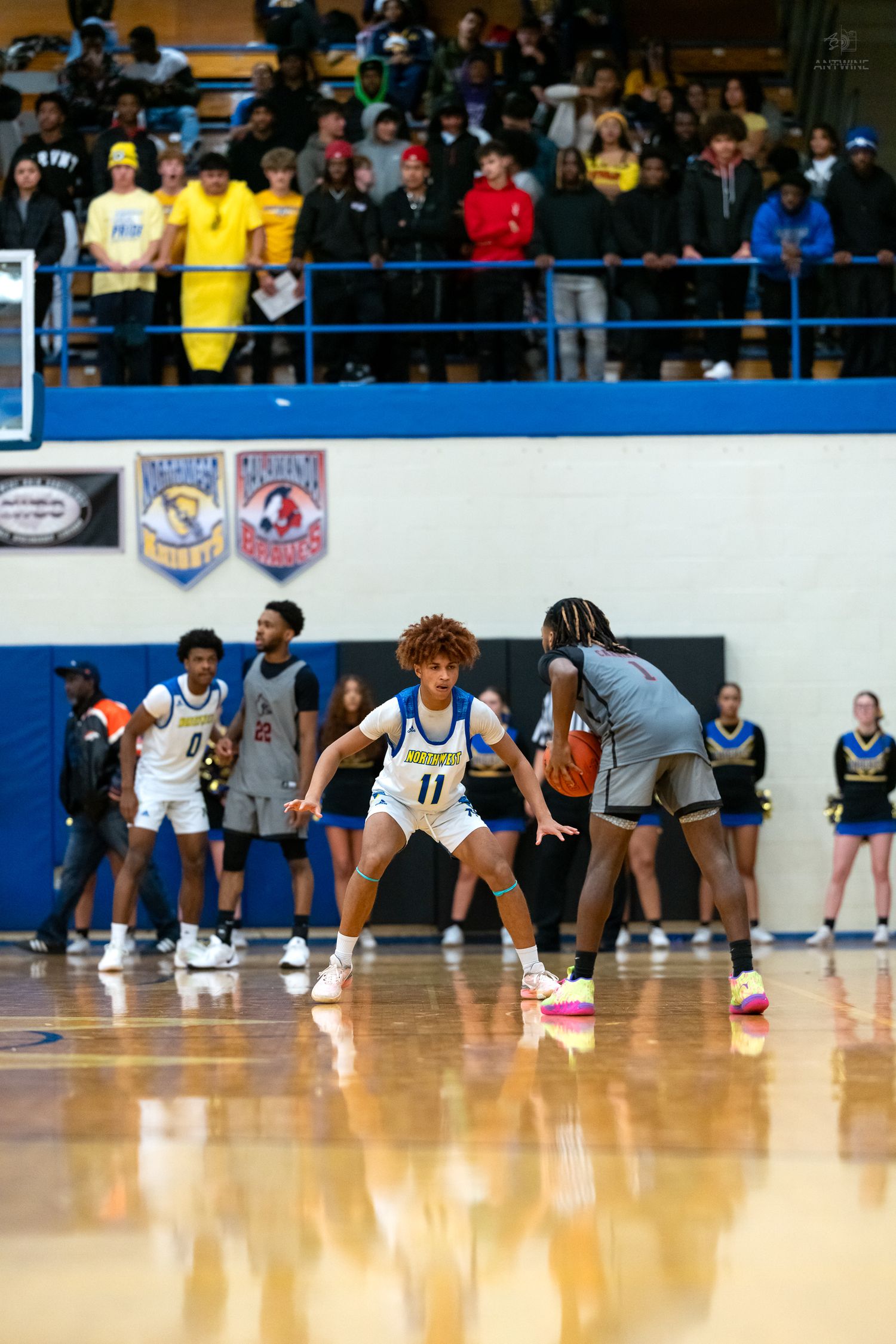 Player #11 in white uniform plays defense against opponent during crowded high school basketball game.