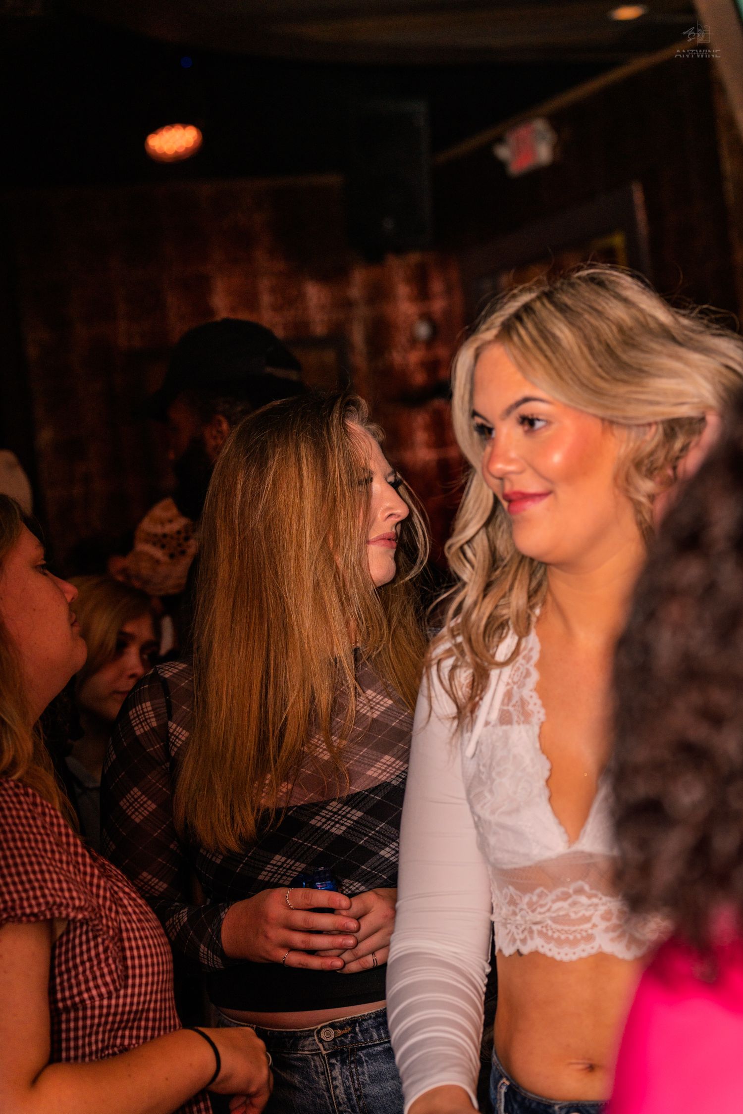 Two young women with long blonde hair socializing at a lively indoor bar or nightclub event.