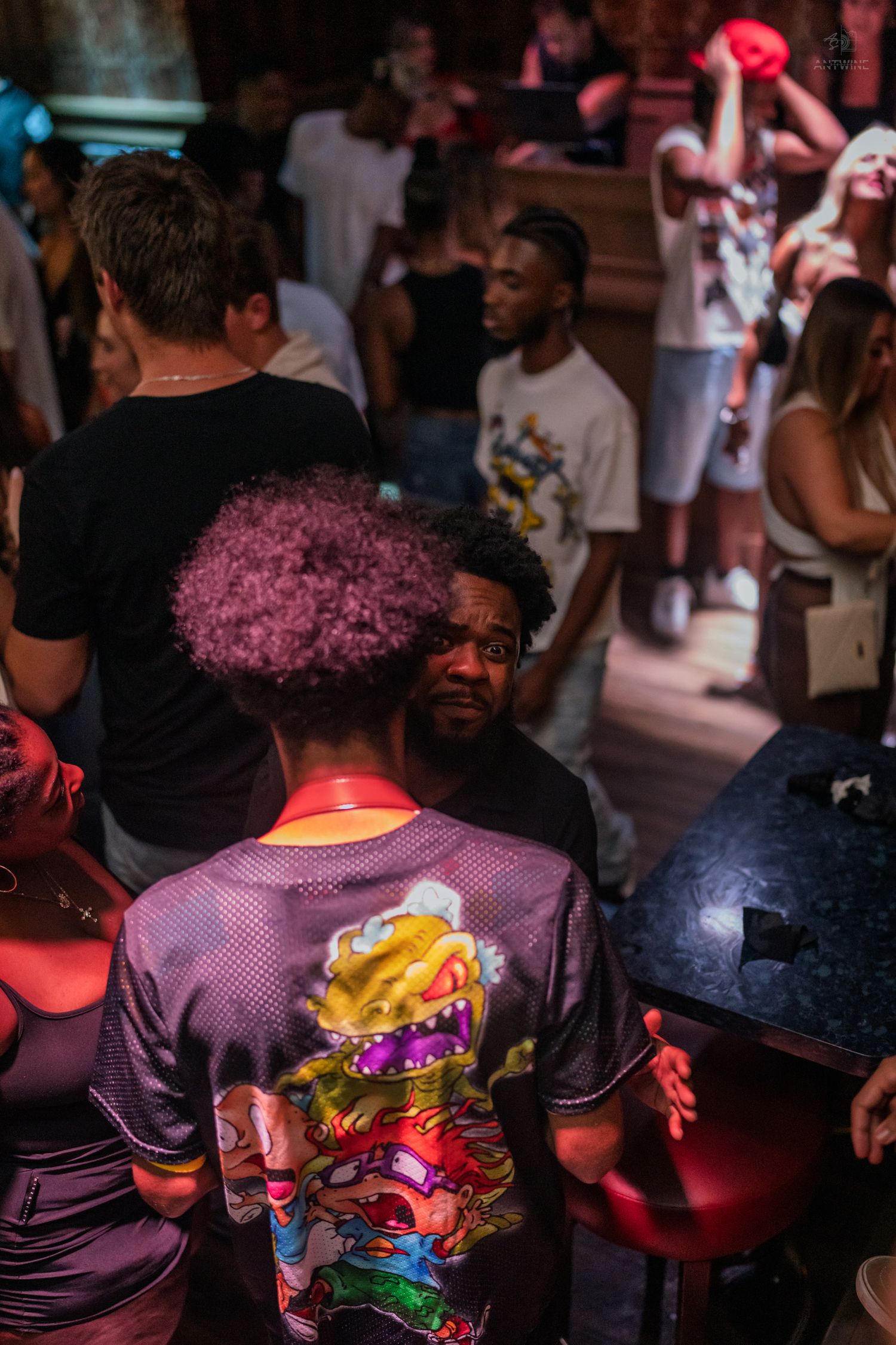 Person wearing colorful graphic tee with purple hair stands in a crowded nightclub under warm lights.