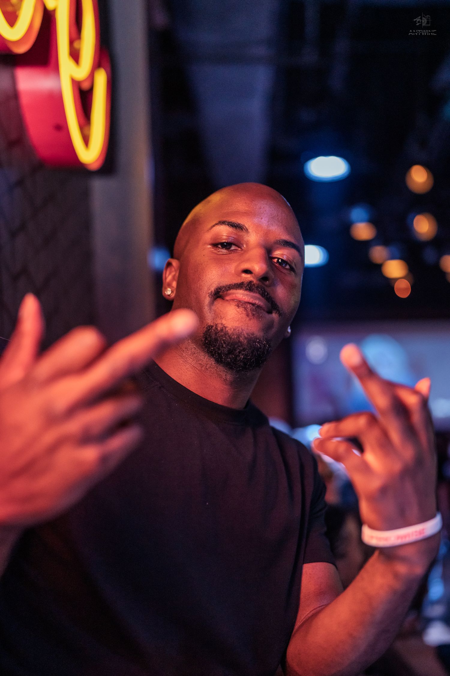 Bald Black man in black t-shirt poses confidently with hand gestures under neon lights at a nightclub.
