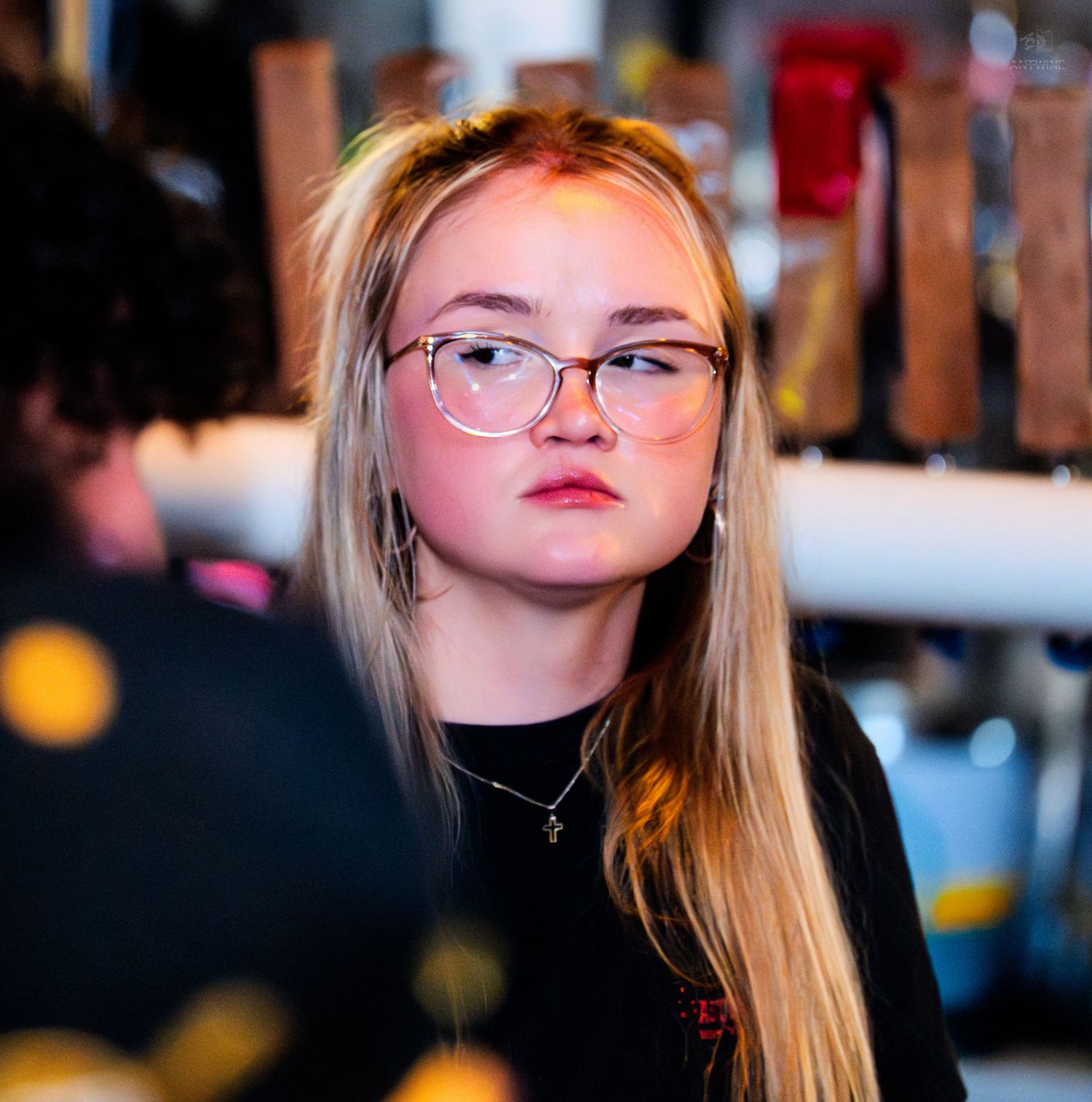 Young blonde woman with glasses and necklace at a busy bar or nightclub event.