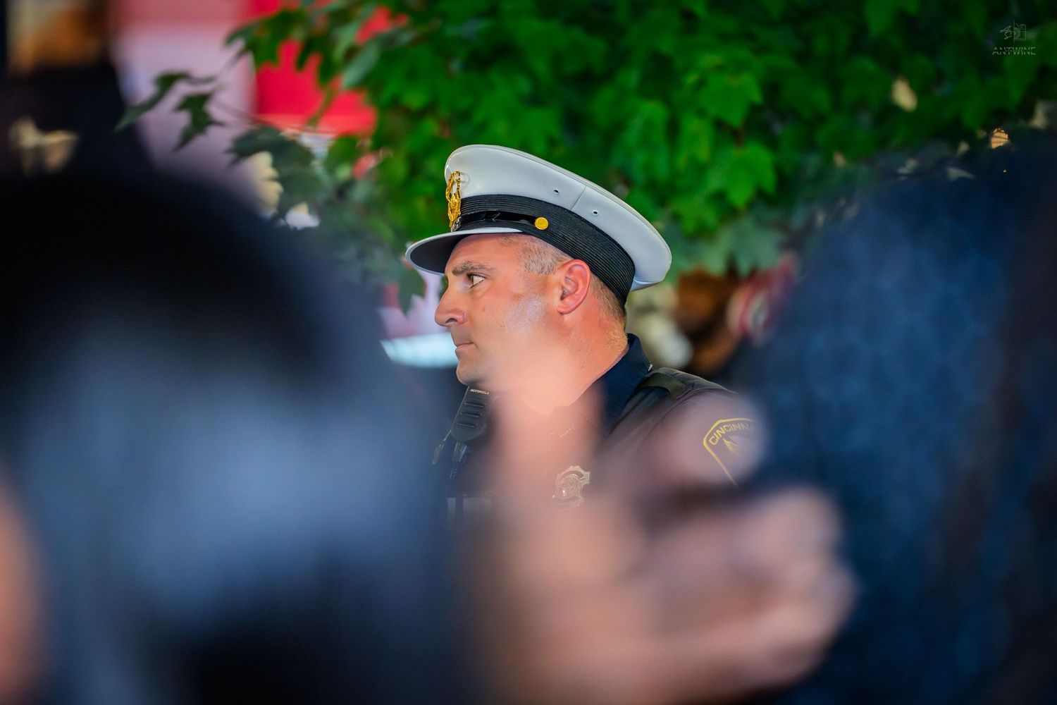A police officer in a white cap and dark uniform stands alert among a crowd outdoors.