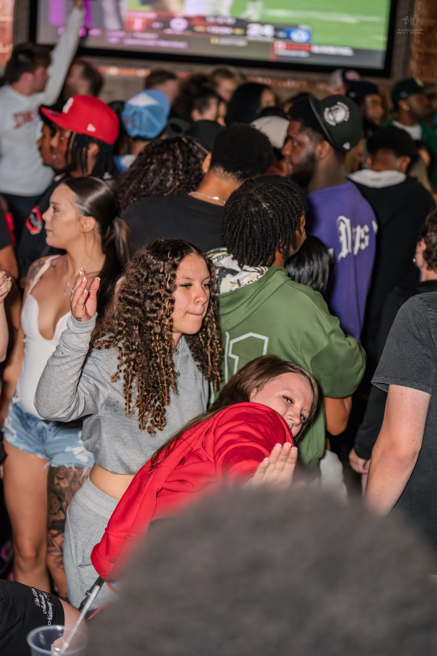 Diverse crowd of young adults gathered in a packed sports bar watching a game on a big screen.