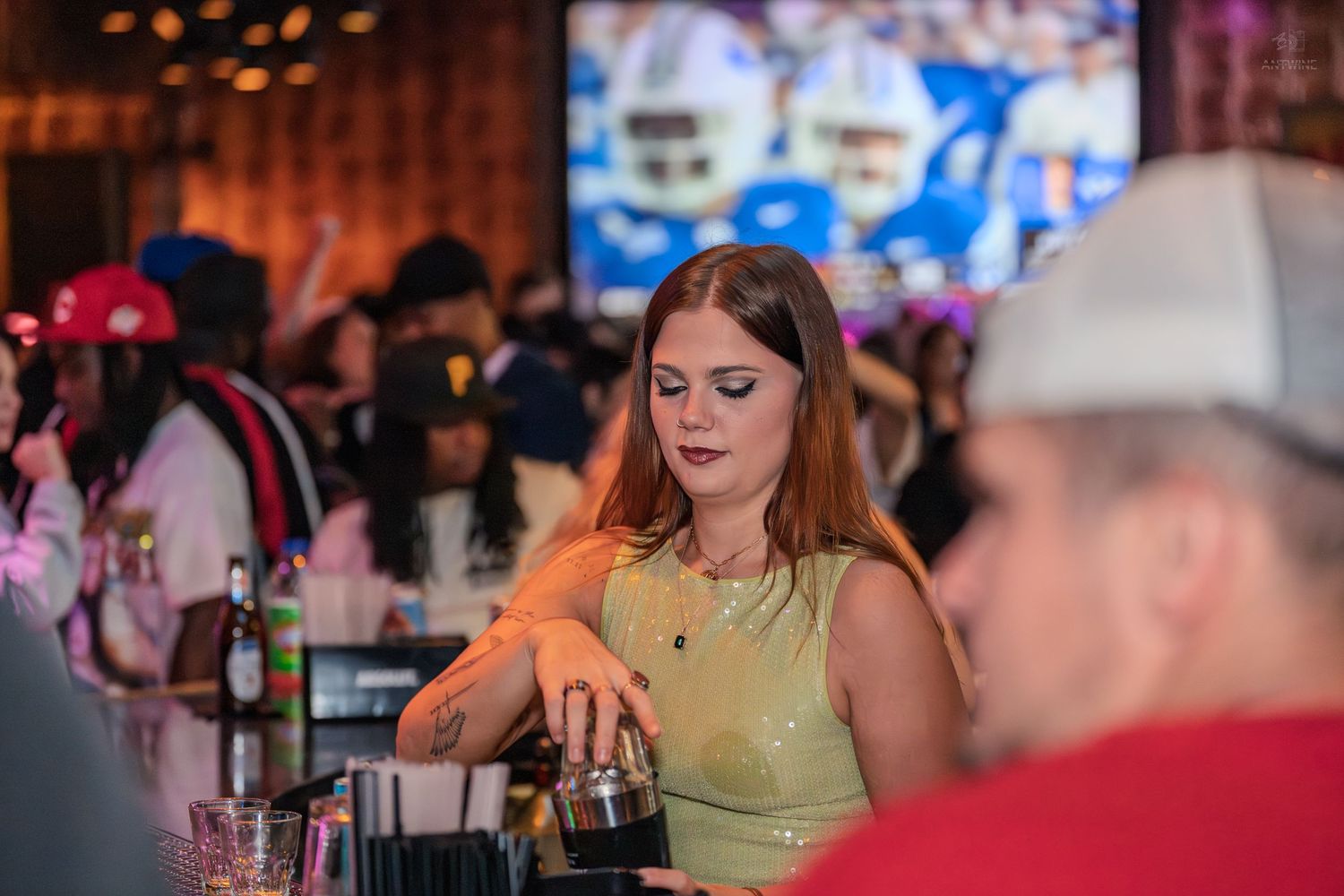 Female bartender in green top mixing drinks at a busy sports bar with TV in background.