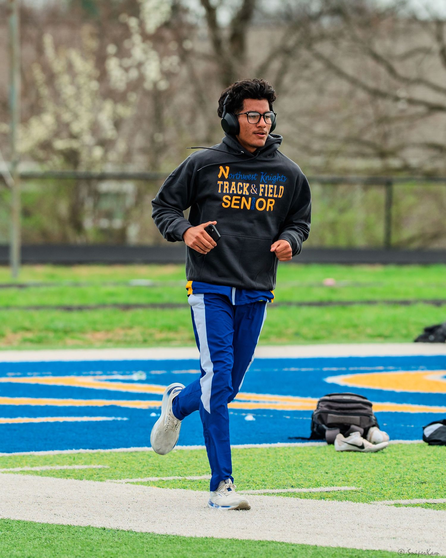 Male track and field senior warms up in black hoodie and blue pants at outdoor track.