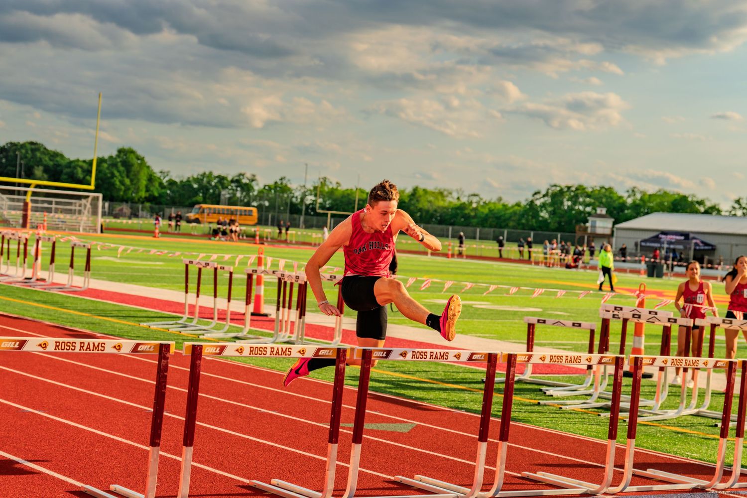 Male athlete leaps over hurdles on red track during outdoor meet.