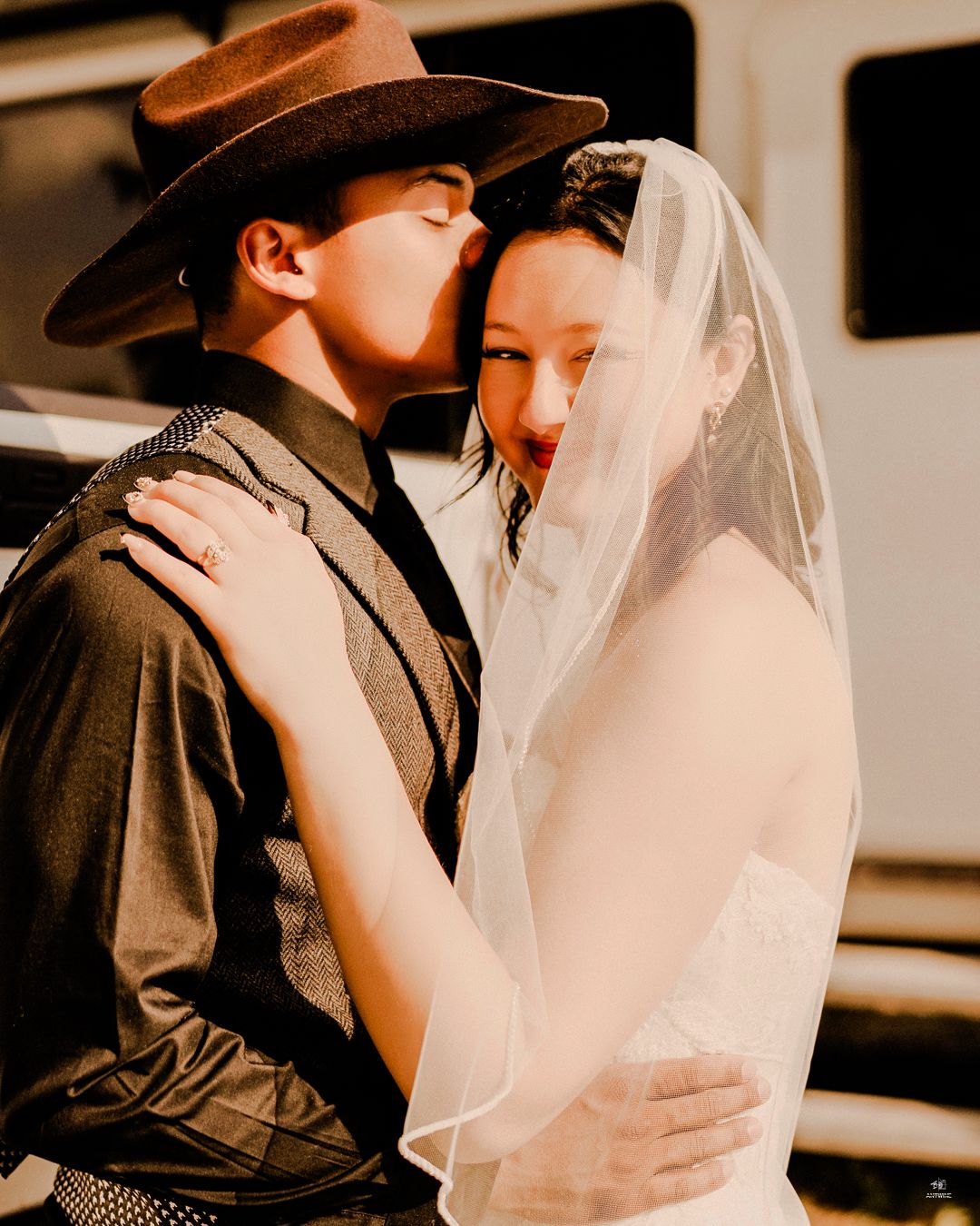 Groom in a cowboy hat kisses the bride's cheek as she smiles joyfully during their wedding celebration.