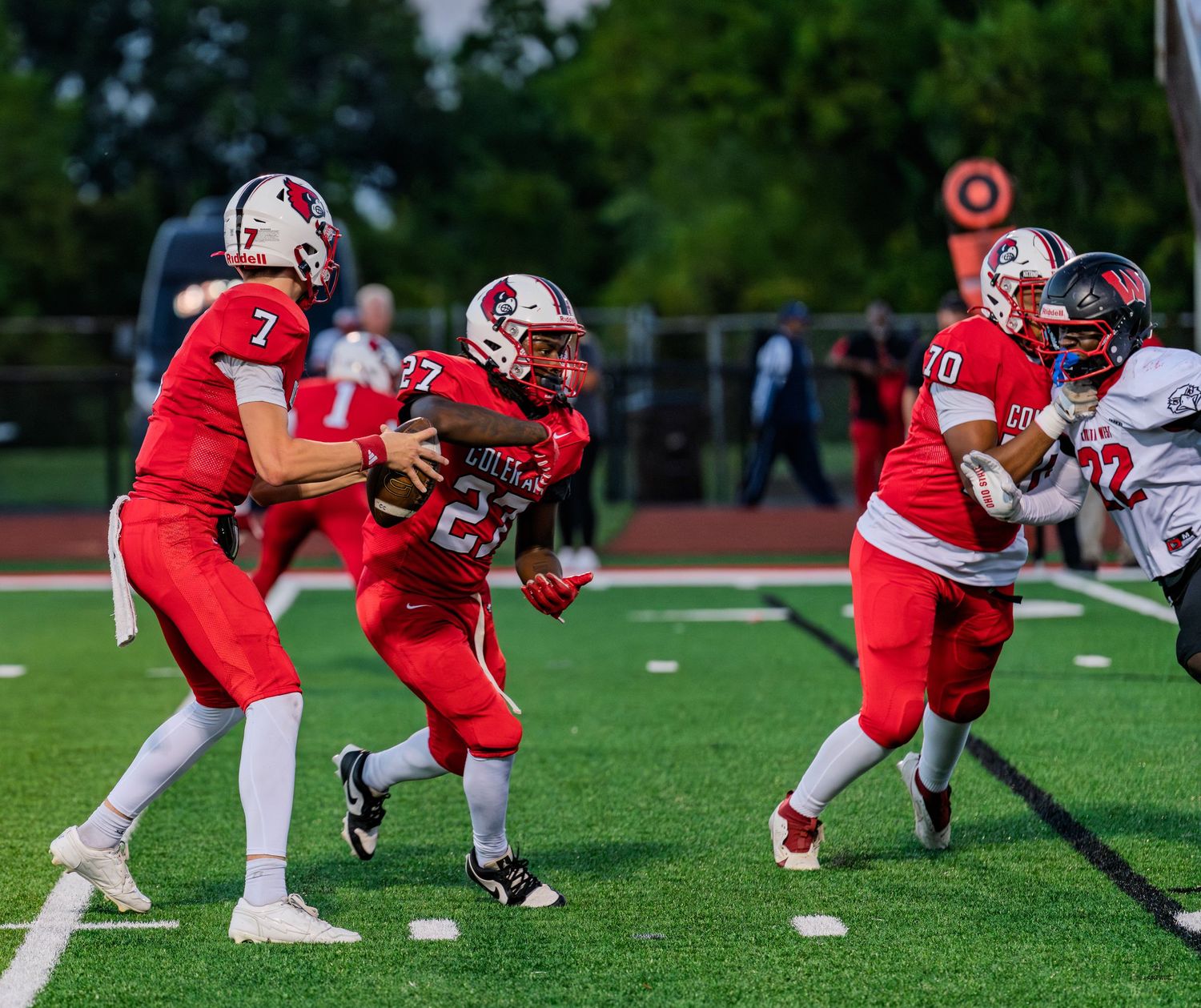 Red-uniformed quarterback number 7 hands off the ball to running back number 27 during a football play.