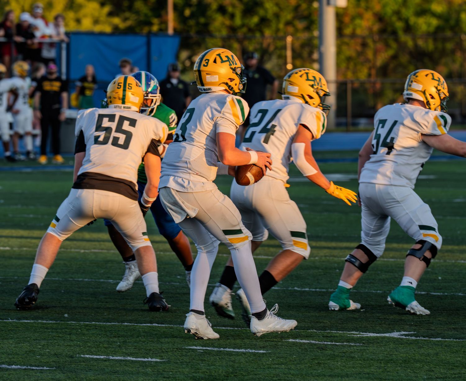 High school football players in gold uniforms execute a play during a sunny evening game.