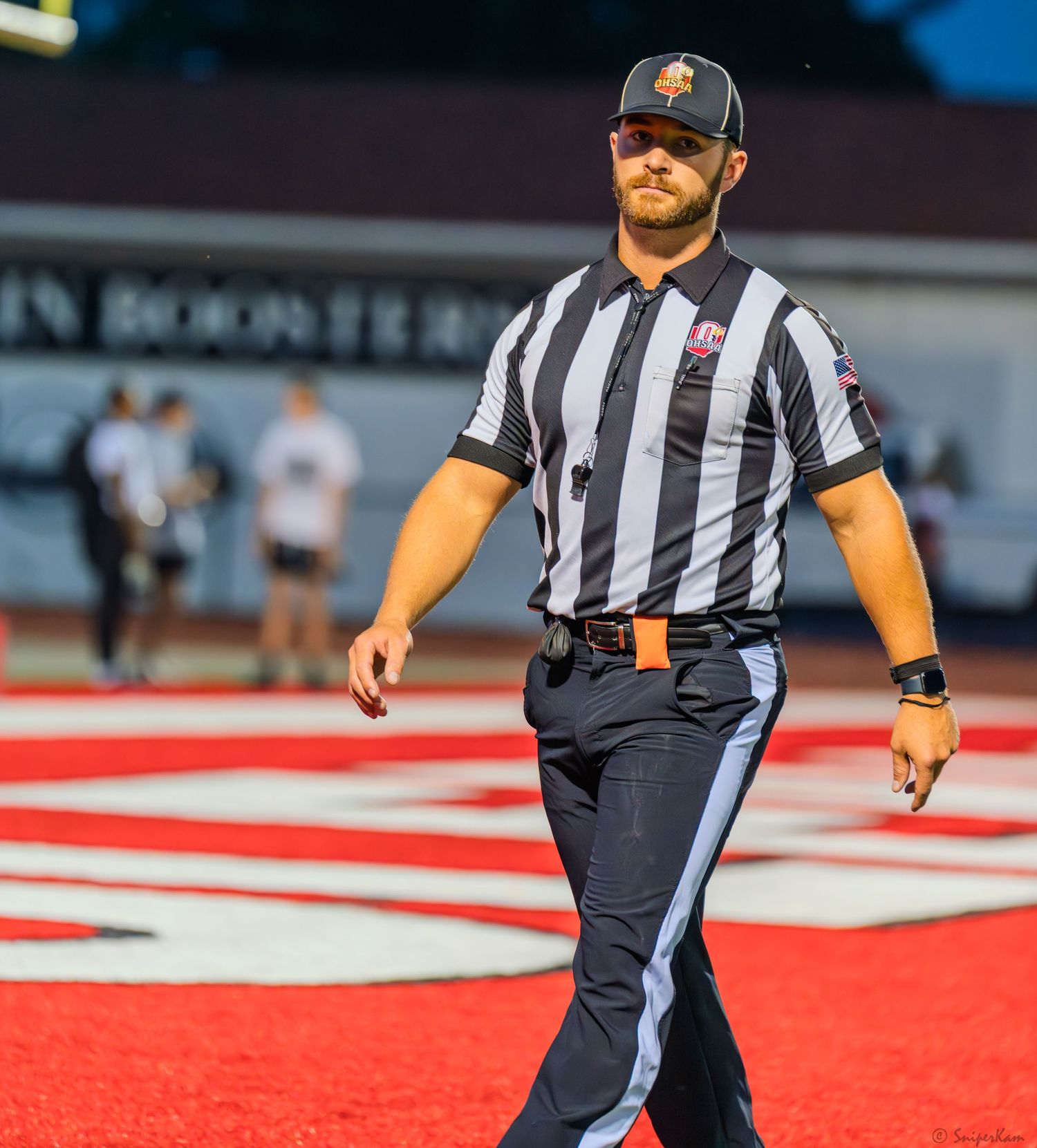 Football referee in black and white stripes walks confidently across red turf during a night game.