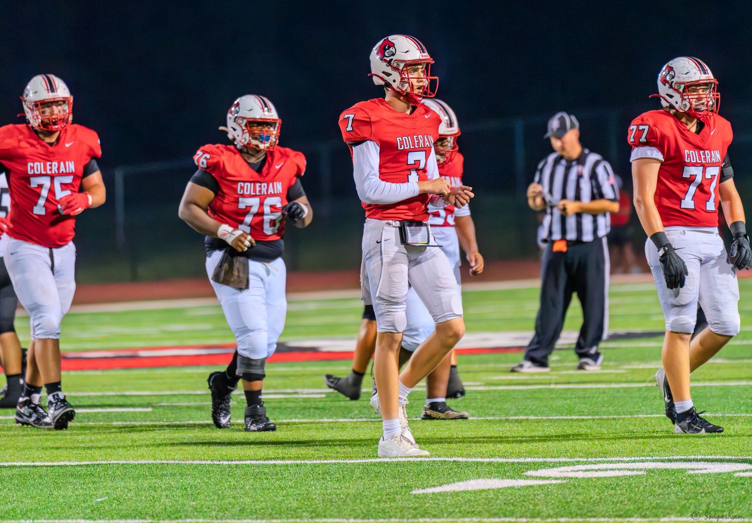 High school football players in red Colerain jerseys walk on the field during a night game.