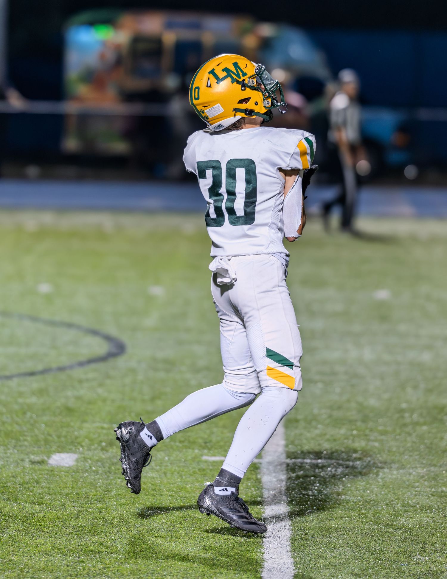 Youth football player #30 in white and gold uniform jogs along the sideline during a night game.