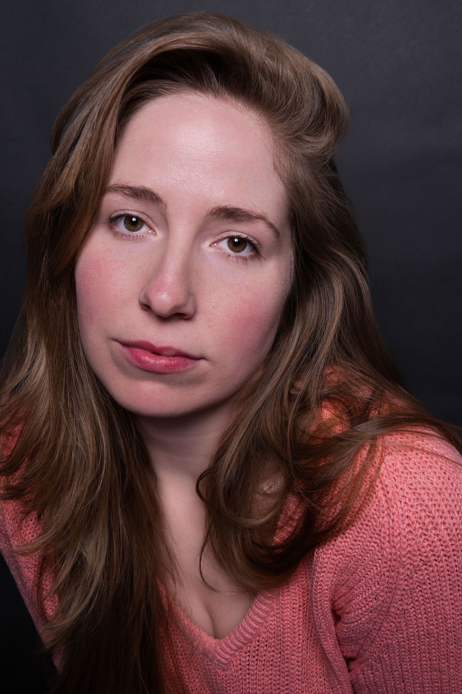 Close-up studio portrait of a young woman with long brown hair wearing a pink knit sweater.
