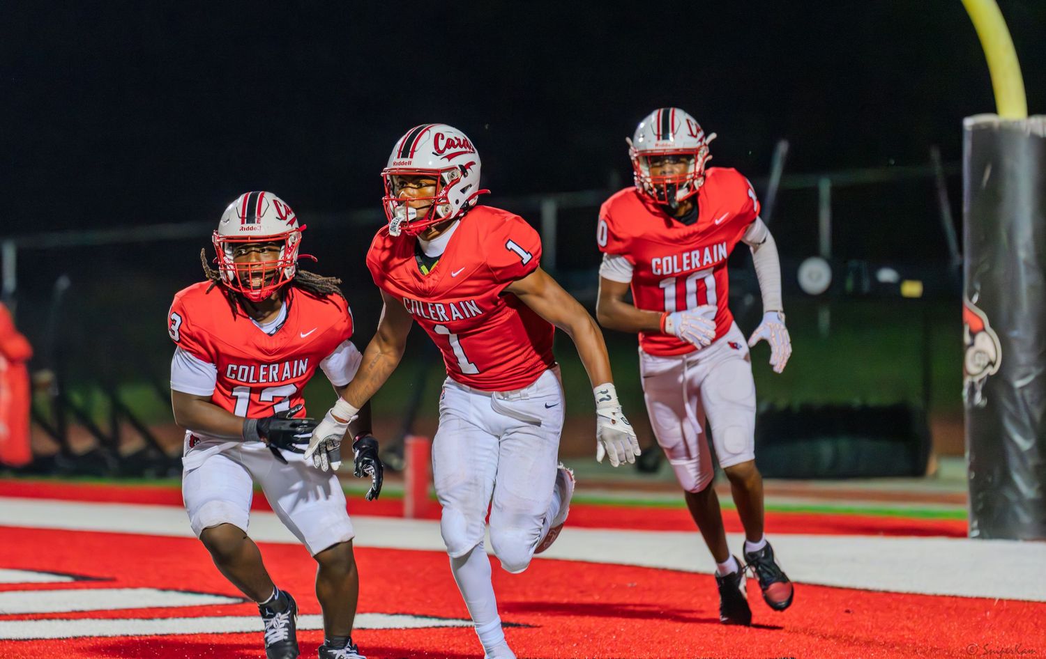 Three Colerain football players in red uniforms celebrate together after a touchdown play.