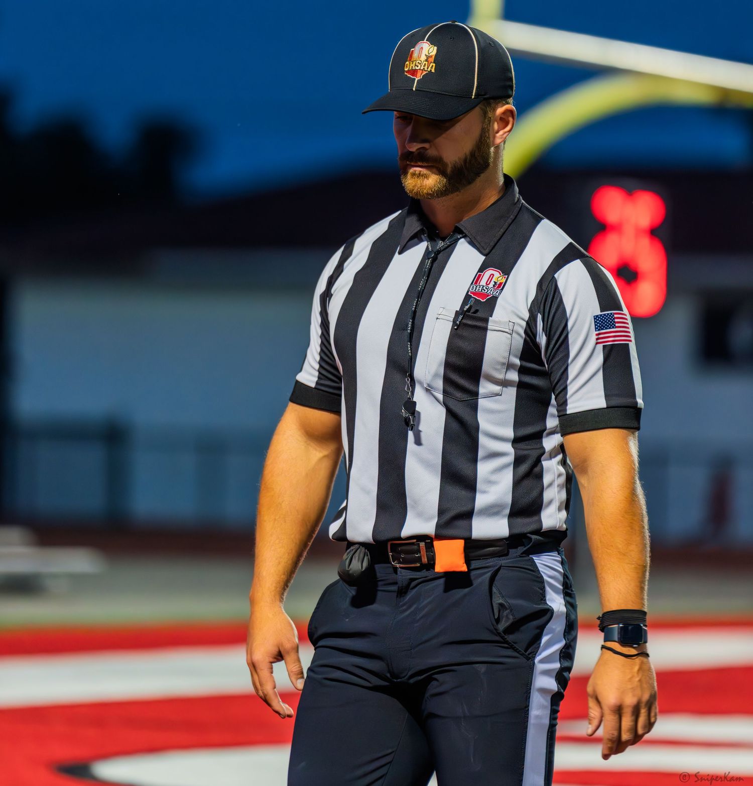 Football referee in black and white striped uniform stands on red turf field during a night game.