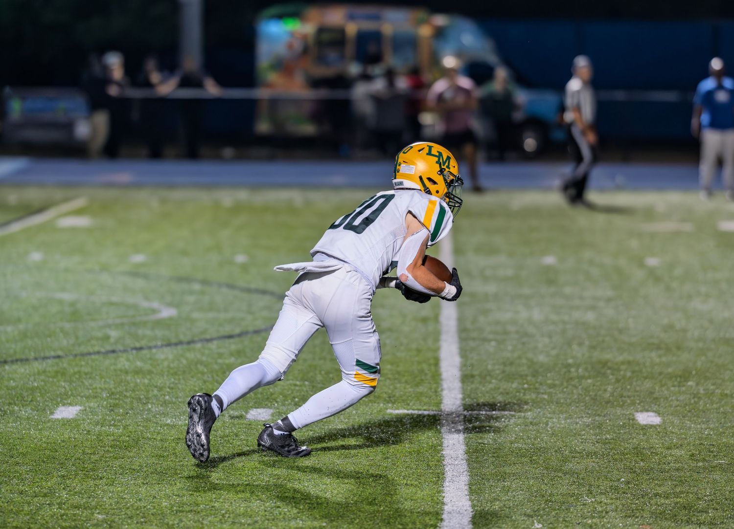 Youth football player in green and white uniform runs with the ball along the sideline during a night game.