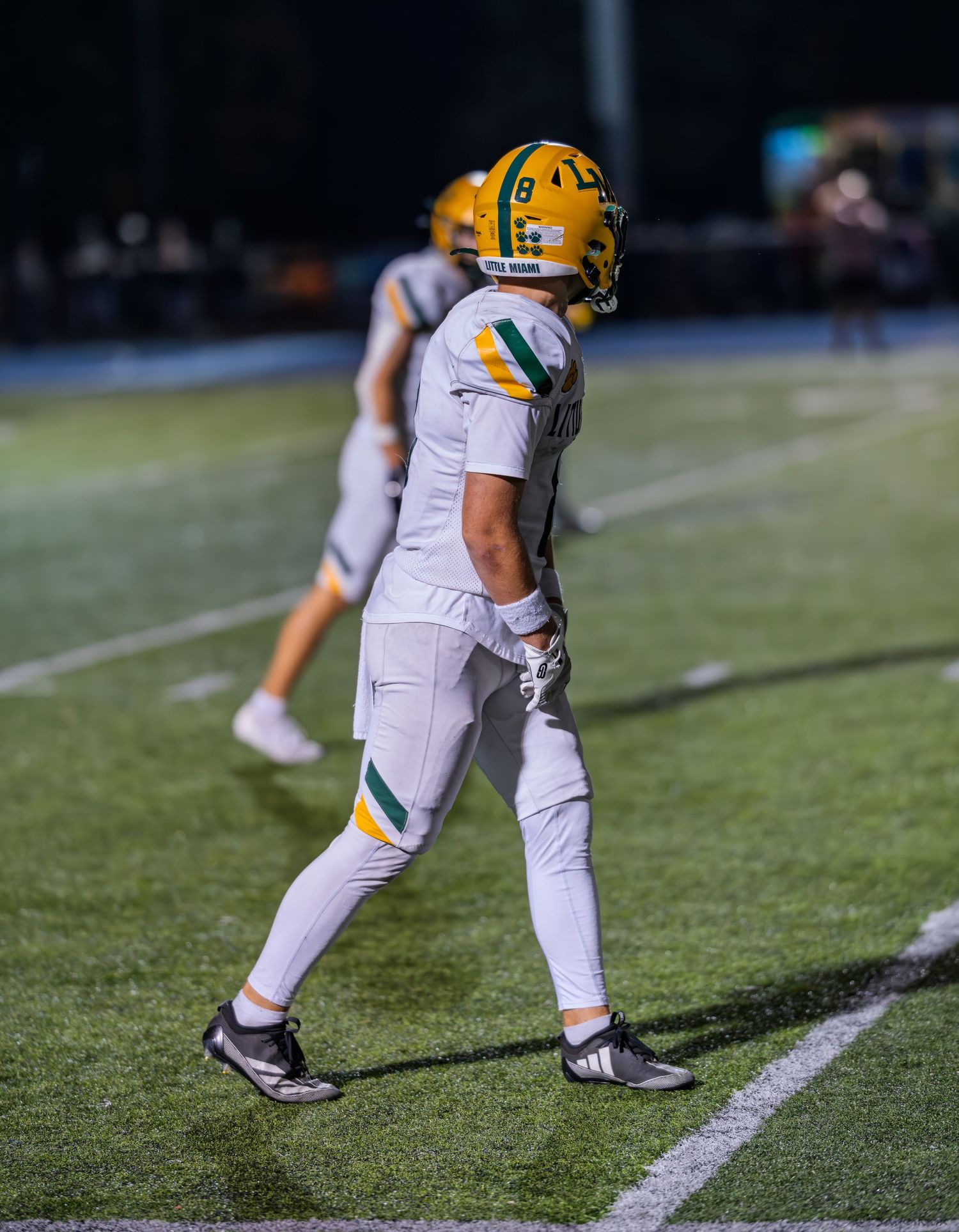 Football player in white and green uniform walks along sideline during night game.