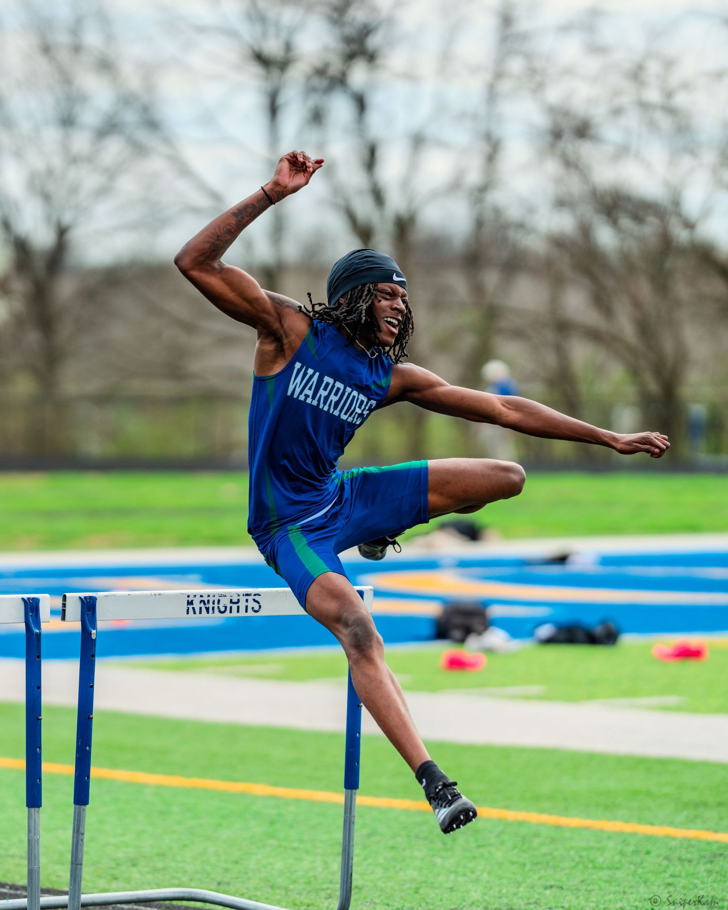 Male Warriors athlete leaps dramatically over hurdle at Knights outdoor track and field meet.