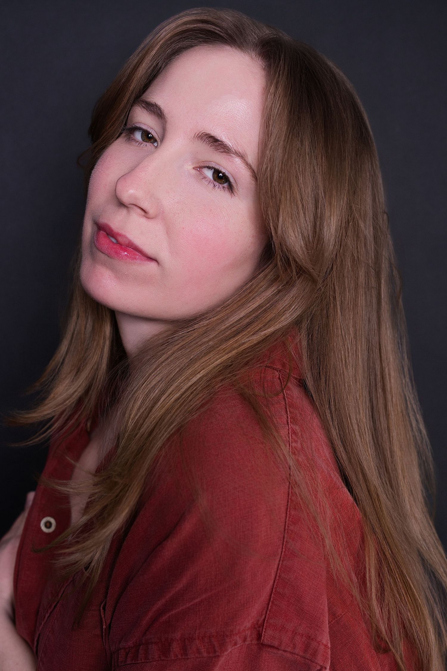 Young woman with long hair in red shirt looking over shoulder with subtle expression against gray background.