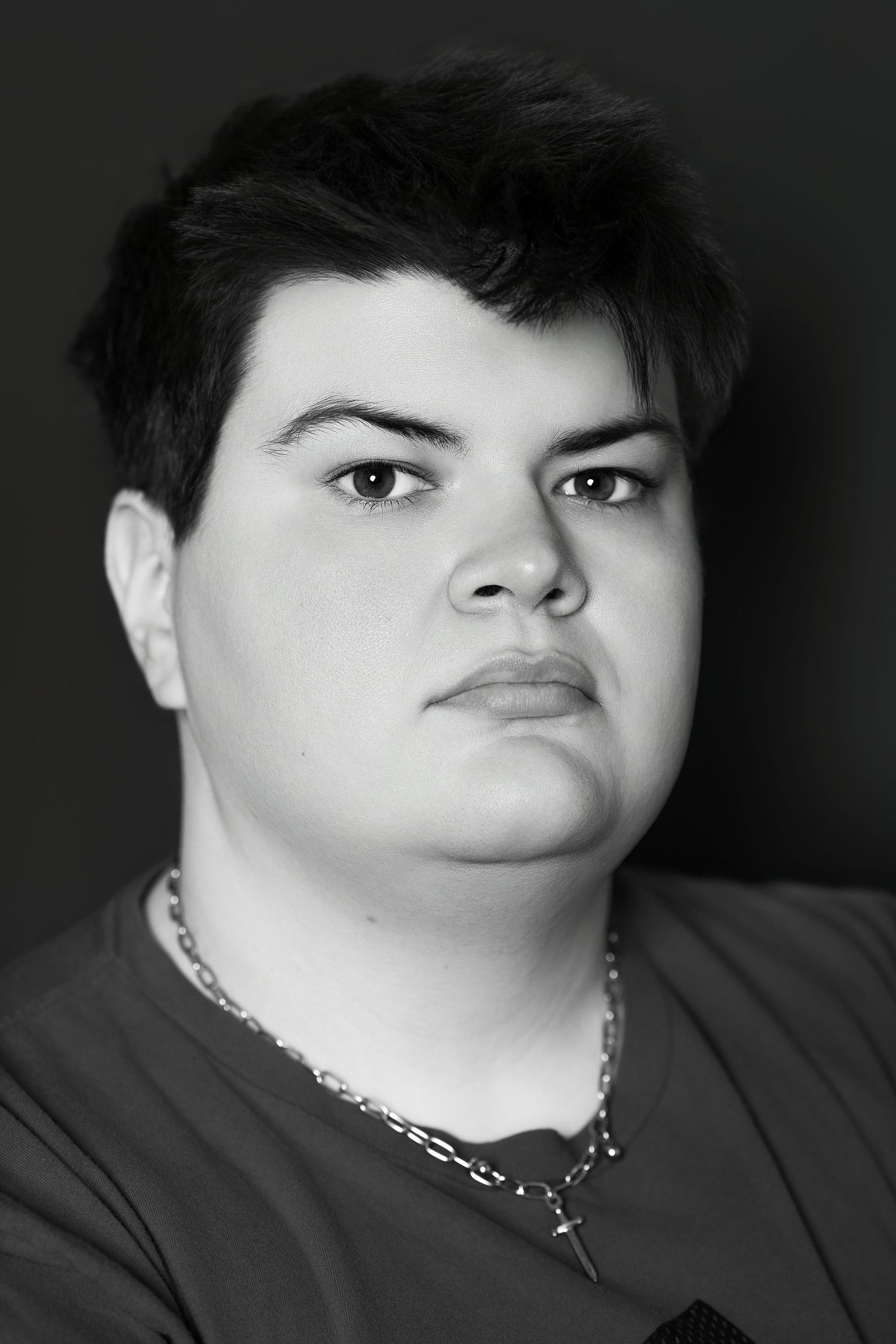 Black and white close-up portrait of a young person wearing a chain necklace with a cross pendant.
