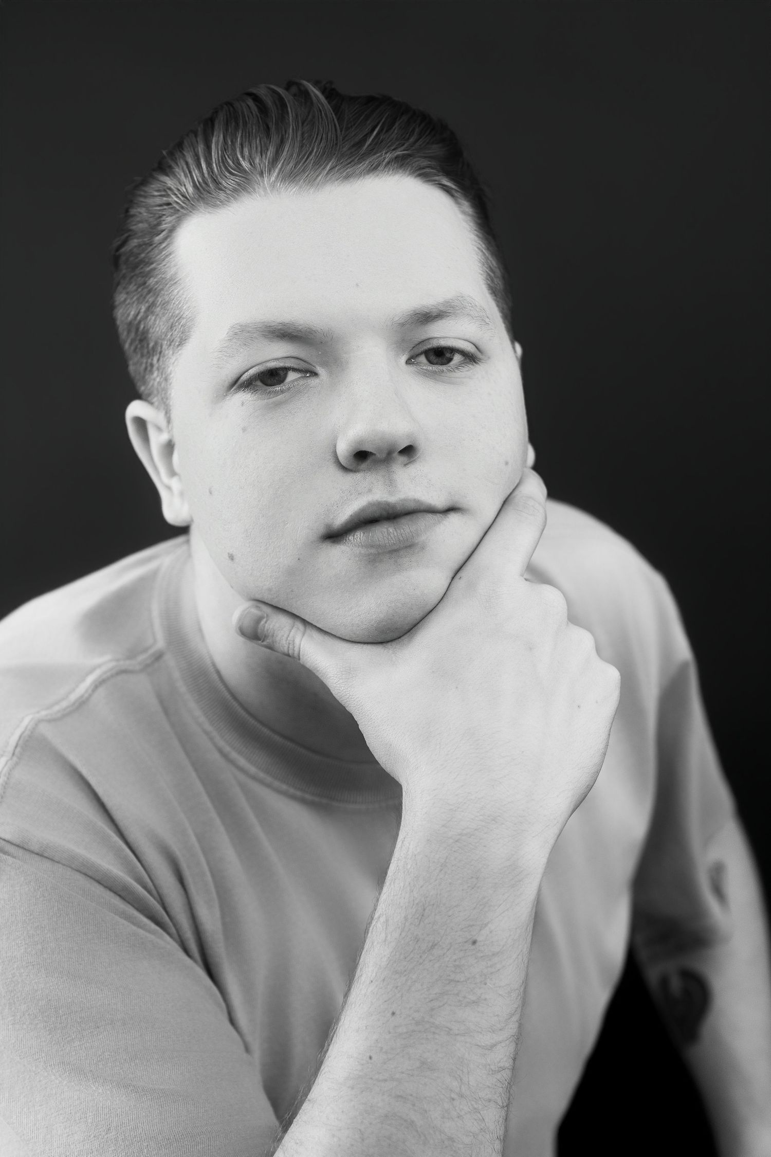 Black and white portrait of young man resting cheek on hand with serious expression on dark background.