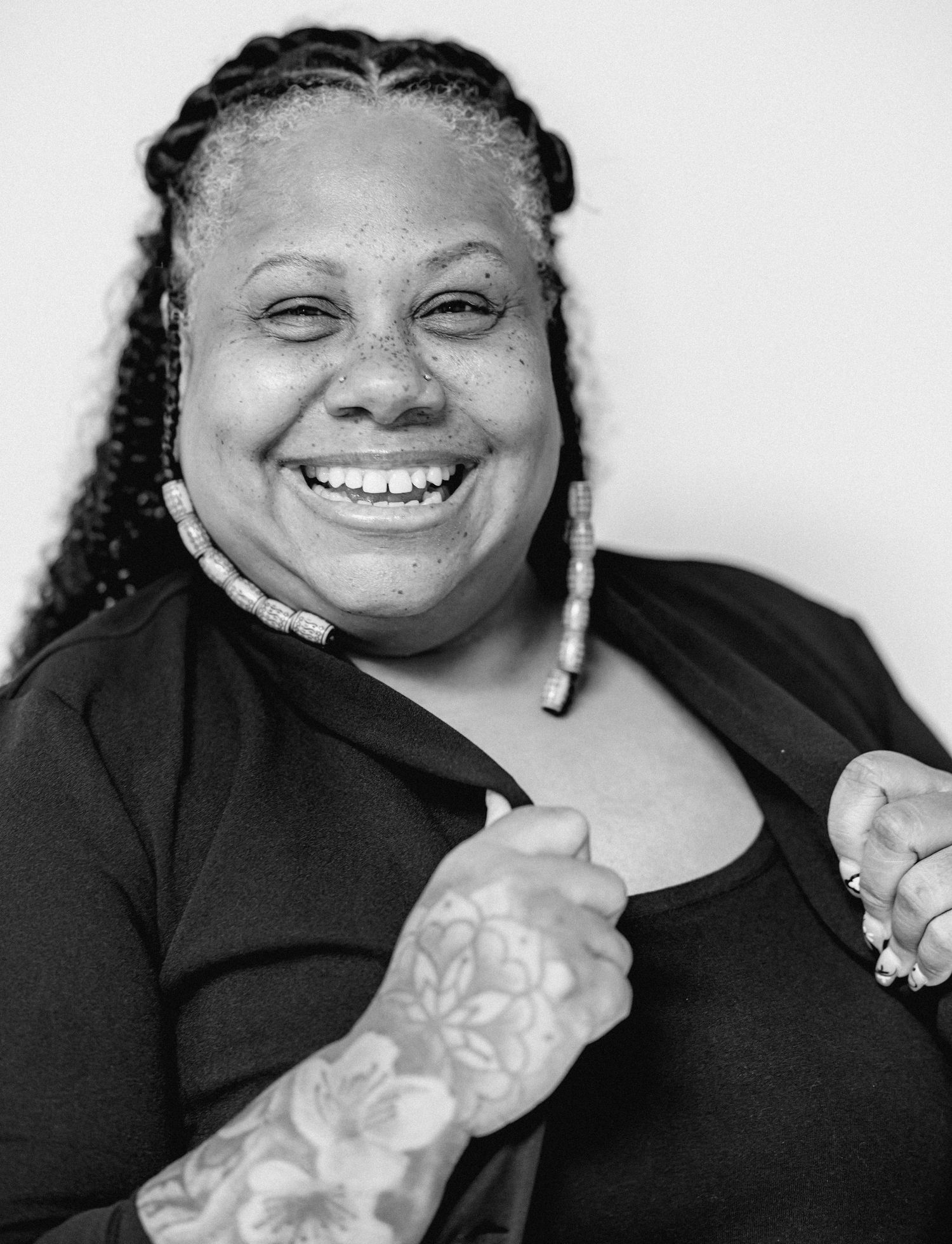 A Black woman with braided hair laughs joyfully, wearing beaded earrings and showing floral arm tattoos in black and white.