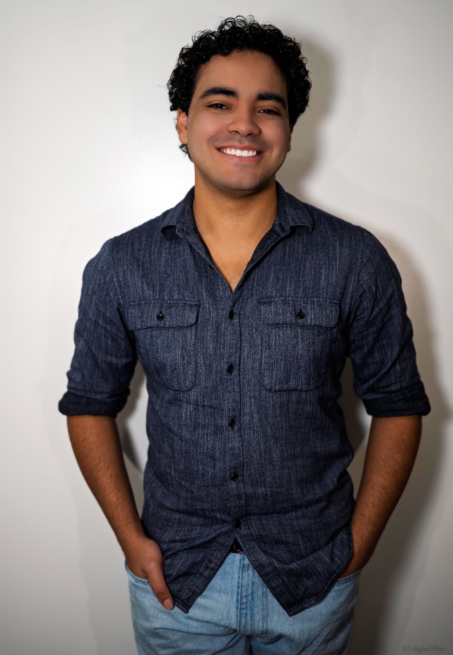 Smiling young man in blue linen shirt with hands in pockets against white wall.
