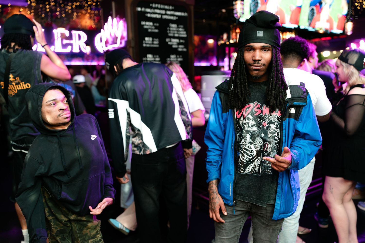 Man in blue jacket and black hat posing at a crowded neon-lit nightclub.