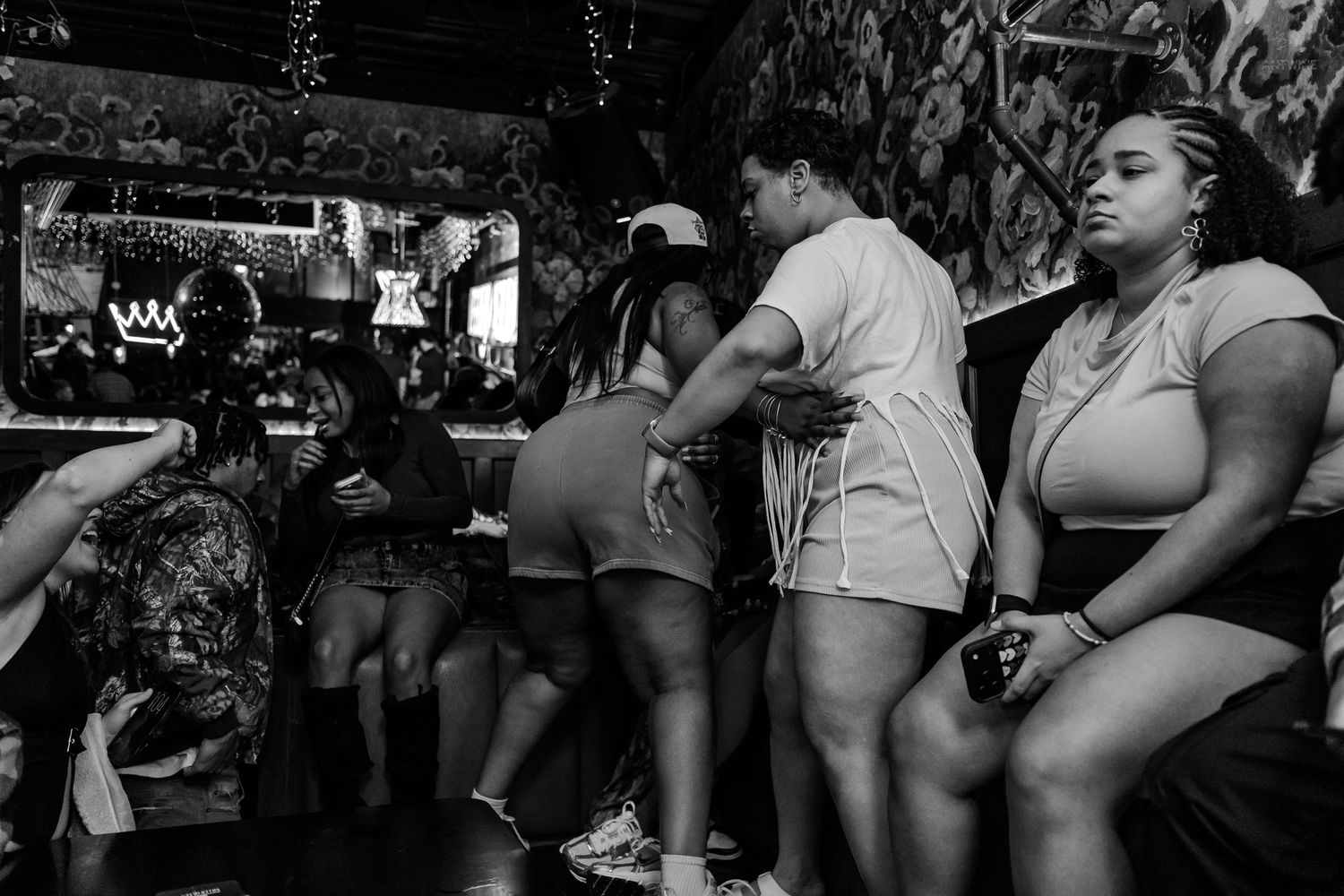 Group of women socializing at a dimly lit bar, some standing and dancing in black and white photography.