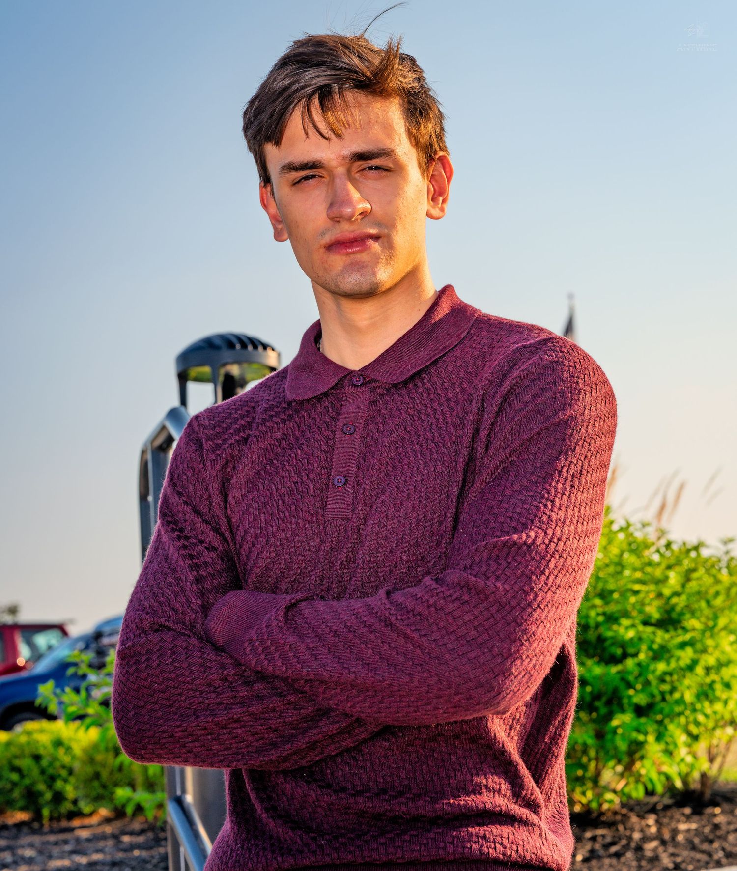 Young man in burgundy knit polo sweater standing outdoors with arms crossed against a blue sky.