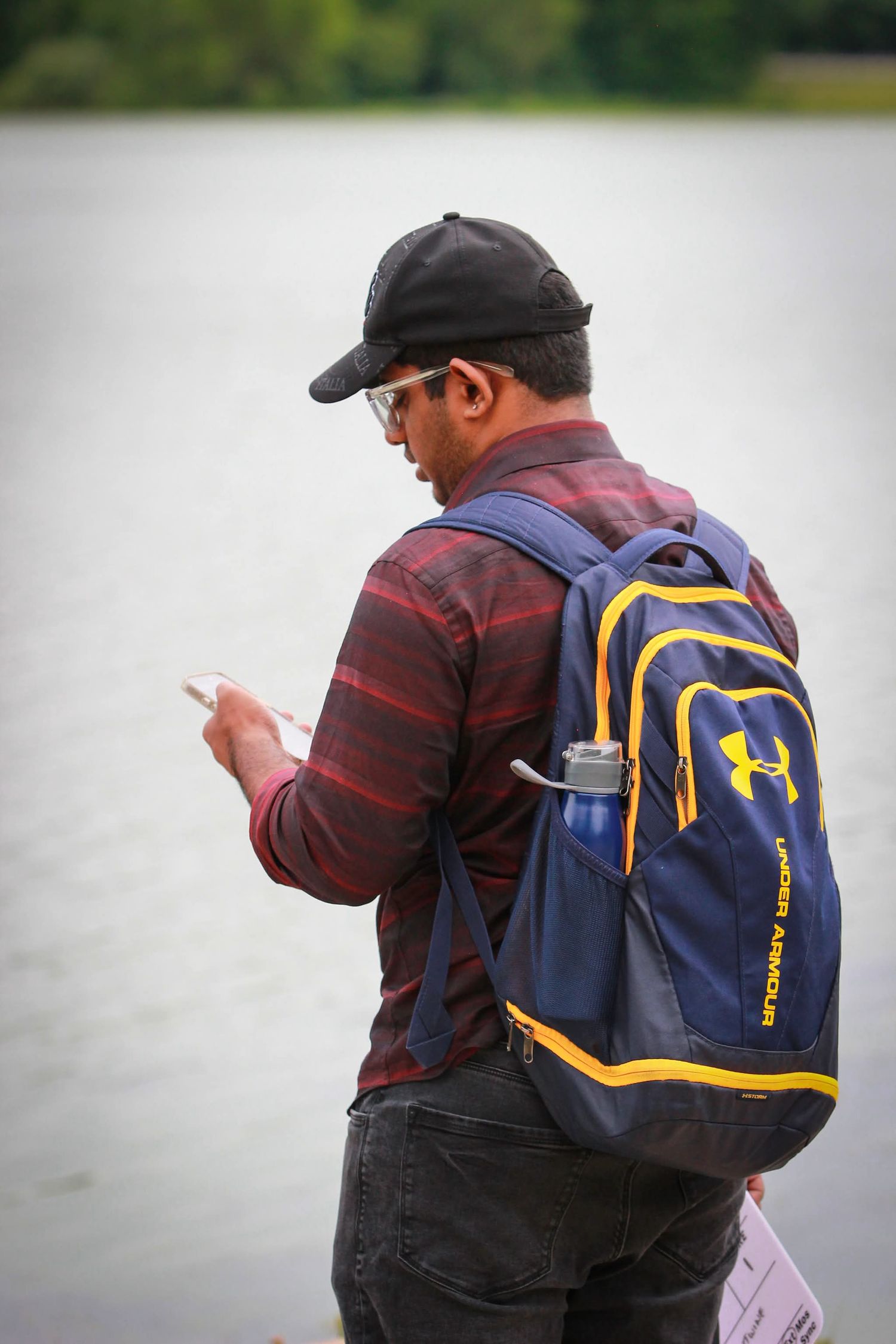 Man wearing Under Armour backpack and plaid shirt checks phone near waterfront.