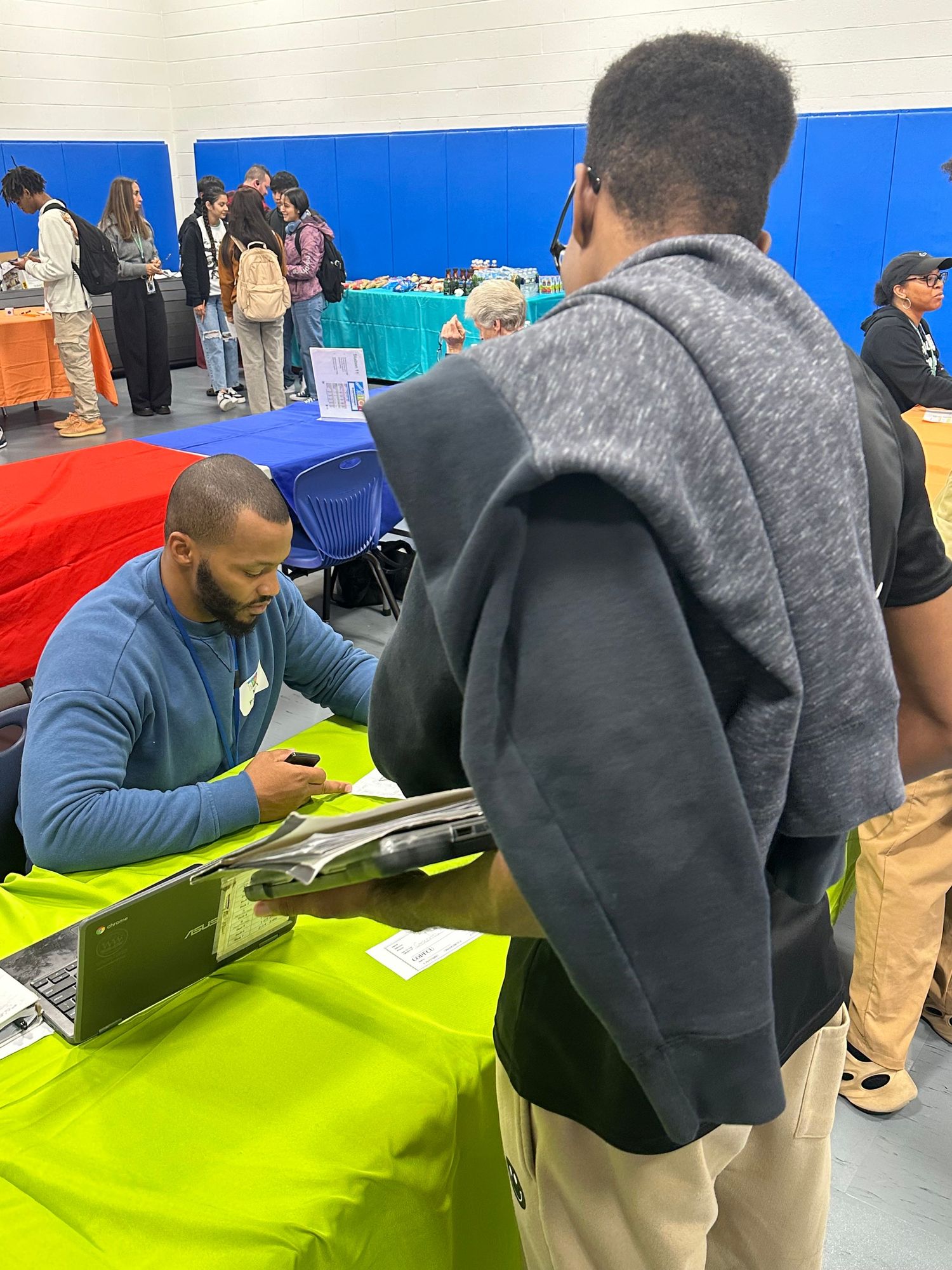 A man signs autographs at a green table during an indoor career fair event in a gymnasium.