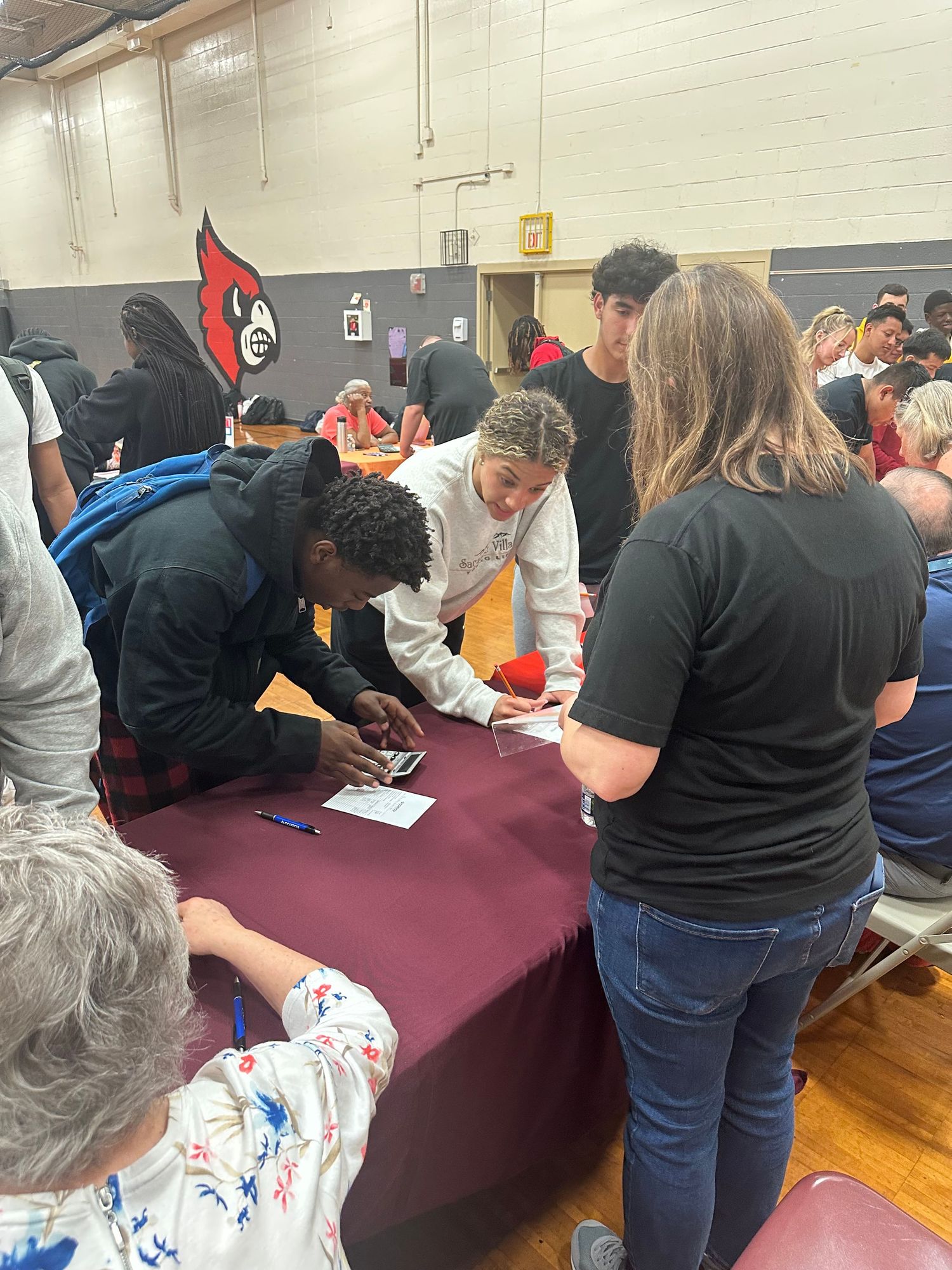 Students signing documents at a maroon table in a school gymnasium during a registration or enrollment event.