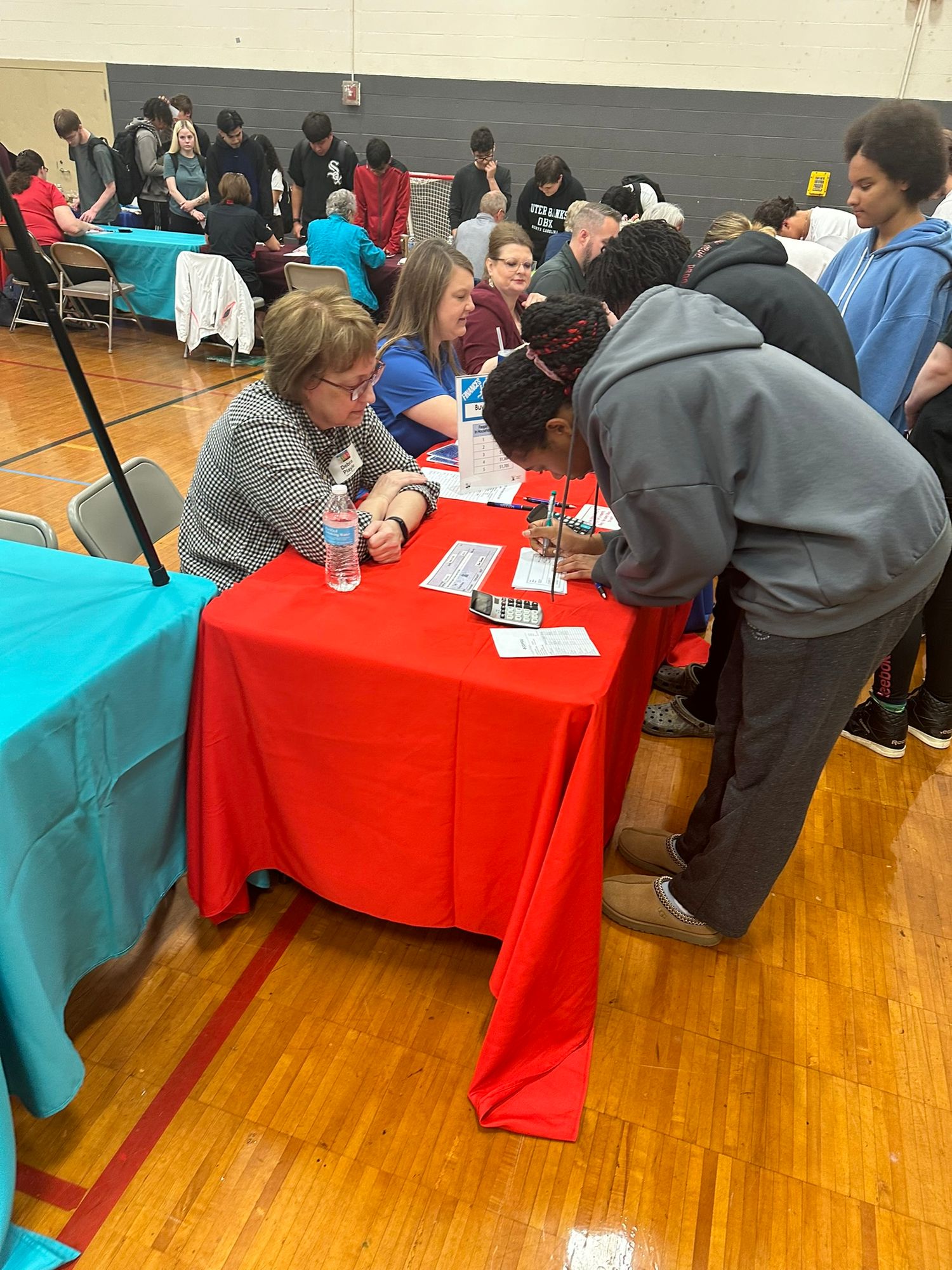 Students interact with recruiters at a school career fair in a gymnasium with colorful tablecloths.