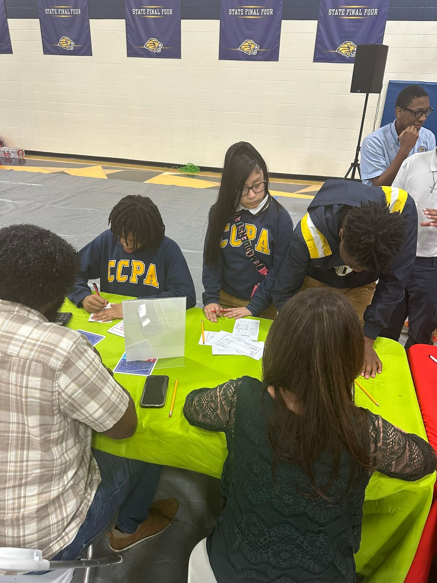 Students in CCPA jackets work at a green table during a State Final Four event in a school gymnasium.