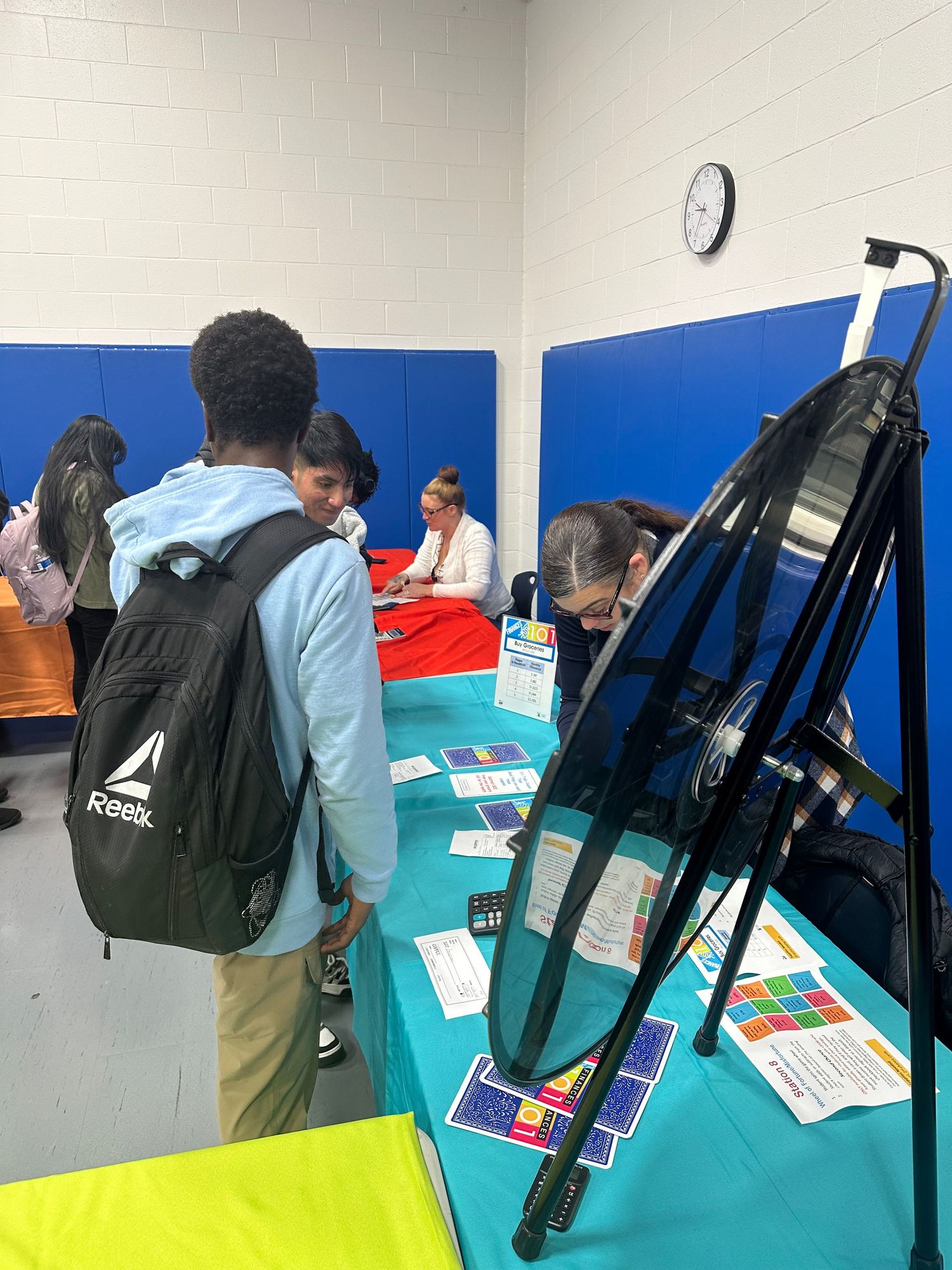 A student with a Reebok backpack browses informational materials at a teal-draped table at a school career fair.
