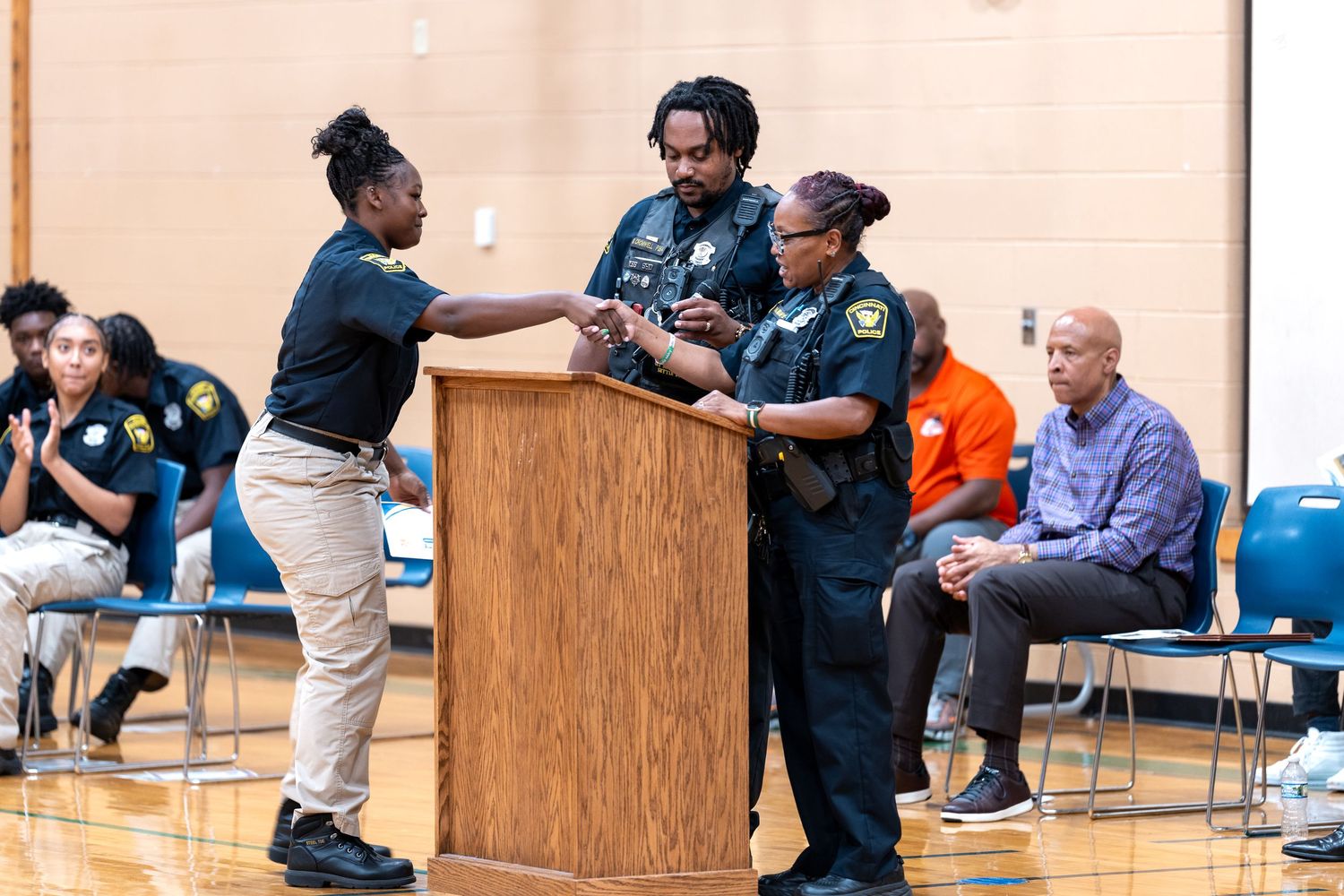 A student receives an award at a podium from two officers during a school ceremony in a gymnasium.